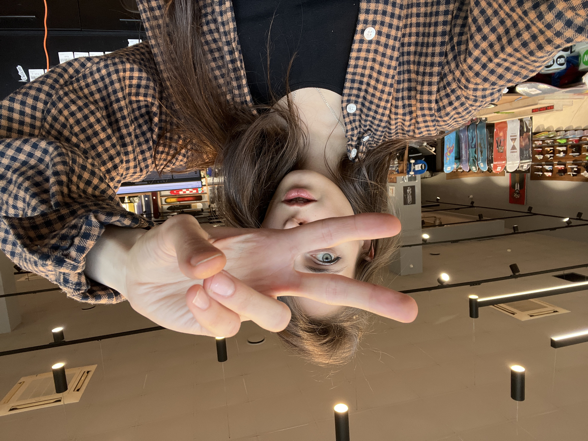 A woman wearing a checkered shirt and a black top makes a peace sign towards the camera. The photo is taken from an upside-down perspective, placing the woman in the foreground. In the background, a retail store with various products displayed on shelves and hanging racks is visible. A row of skateboards is hanging on a wall. The location may be a store in the city of Krasnoyarsk, Russia. Overhead, ceiling-mounted track lighting illuminates the interior space.