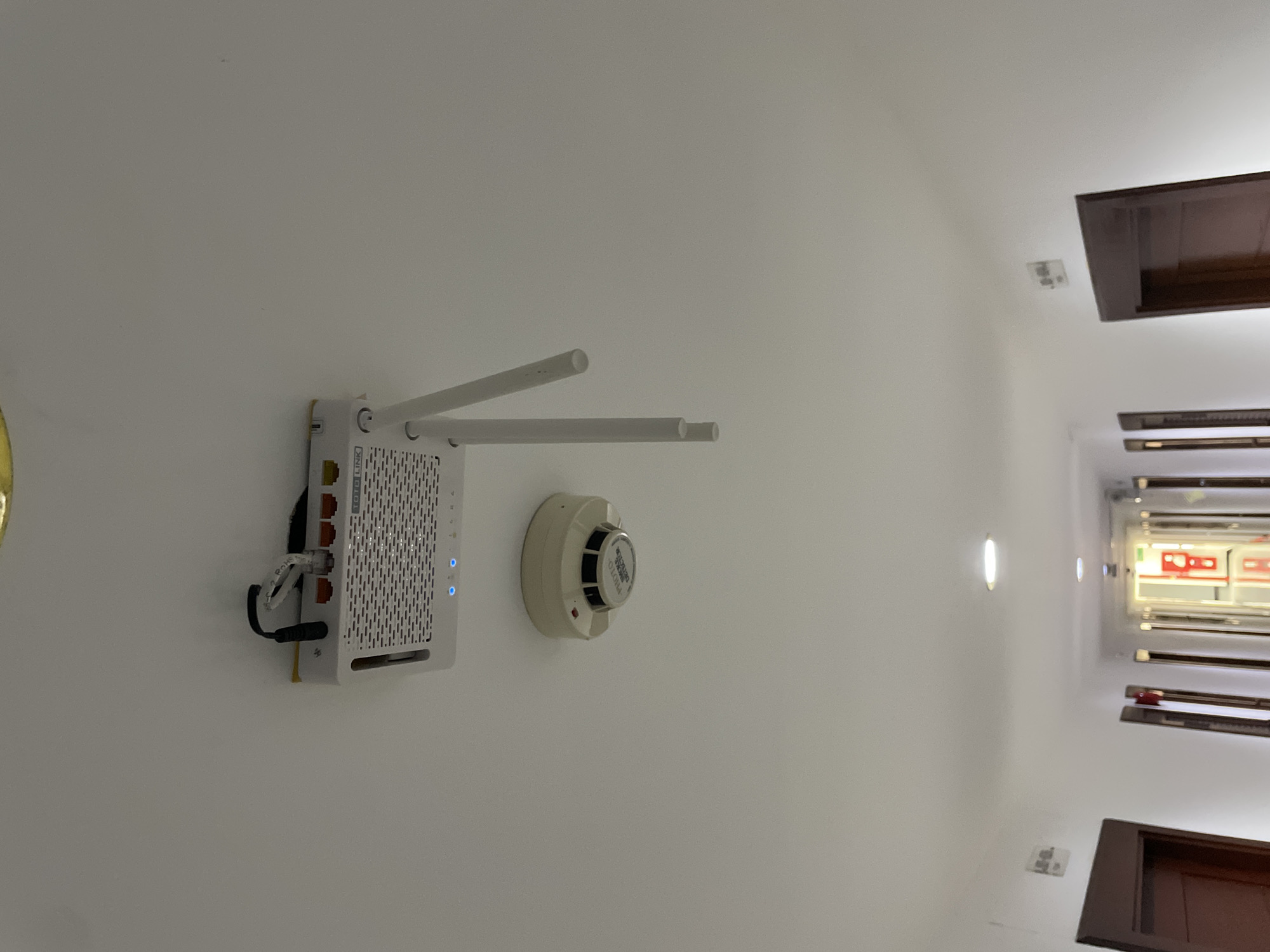A white Wi-Fi router is mounted on a white ceiling in a hotel hallway in Pattaya City, Thailand. The router has two antennas, both extended upwards. Below the router is a white smoke detector. The hallway is lined with closed doors on both sides and illuminated by ceiling lights. The floor appears to be tiled.