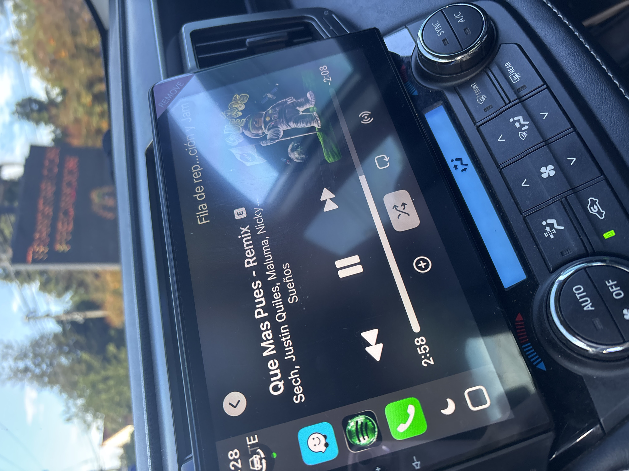 A close-up photograph taken inside a vehicle shows the car's infotainment system screen displaying music playback. The song is "Que Mas Pues - Remix" by Sech, Justin Quiles, Maluma, and Nicky, playing on the music app. The screen indicates the song is 2 minutes and 58 seconds into playback, with 2 minutes and 8 seconds remaining. Several app icons appear at the bottom of the screen, and the vehicle's climate control buttons and knobs are visible on the right. The location cannot be determined to be Guarne, Colombia, or any other specific place.