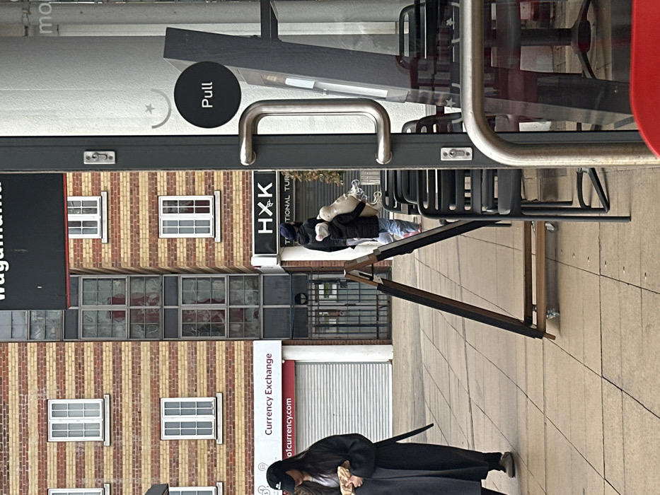 The image displays an urban street scene on Kilburn High Road, London, United Kingdom. In the immediate foreground on the right, the entrance to a glass-fronted establishment is partially visible, featuring a dark grey door frame with a chrome pull handle and a black circular "Pull" sign on the glass. Alongside this, several black metal chairs are stacked or arranged. On the paved sidewalk, a wooden A-frame sign on wheels stands.
Two adults and one child are observed walking from left to right. On the left, a person wearing a black baseball cap, a dark long coat, and light-colored shoes holds a light-brown, paper-wrapped object. To their right, an adult in a dark hooded jacket and a dark blue beanie hat, accompanied by a small child in a dark puffer jacket, carries a large, pale, irregularly shaped item, possibly a plush toy or wrapped object, with a white, fluffy element near the top.
The background features several buildings. To the left, a multi-story building constructed of light and dark brown striped brickwork displays white-framed windows. Below this, a "Currency Exchange" sign is mounted above a closed white roller shutter. In the center, a taller, darker building with a vertical stack of square windows is visible, with a metal gate at ground level. To the right, another striped brick building hosts a shop with a partially visible "HXK Traditional Turkish" sign. A "Wagamama" sign is also partially visible at the top center of the frame.