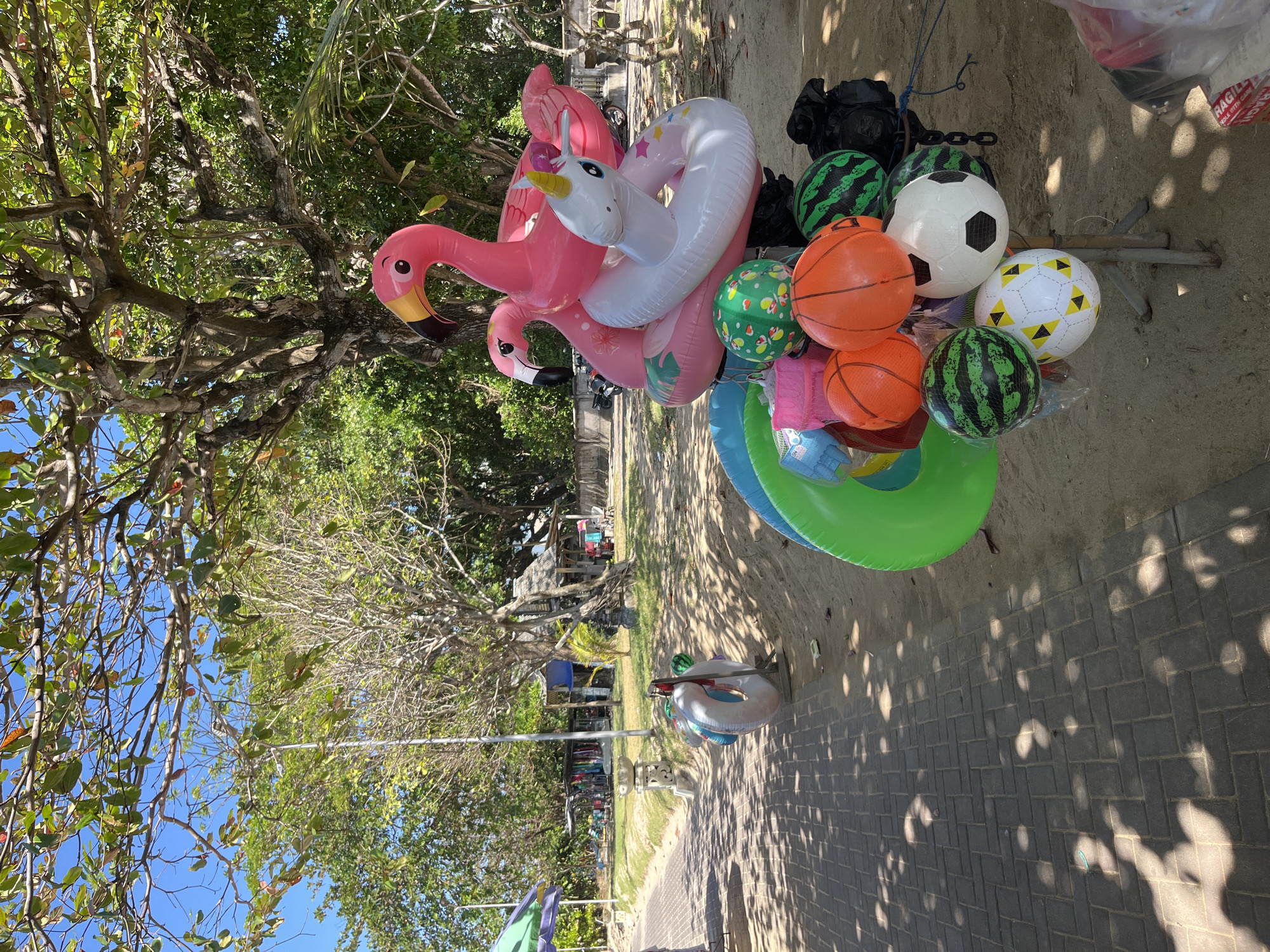The image shows an outdoor scene on a sunny day, likely on a beach in Kuta Selatan, Indonesia, given the presence of typical beach vendors. In the foreground, a display stand holds various inflatable toys and balls. These include a large pink flamingo float, a white unicorn float, green inflatable rings, and several balls designed to look like watermelons, basketballs, and soccer balls. A paved walkway runs along the left side of the image, with sandy ground to the right. In the background, lush green trees provide shade, and further back, there are small structures that appear to be vendor stalls or huts, with some clothing items displayed for sale. A few people are visible in the background, walking near the stalls or near the water's edge, and a motorcycle is parked in the distance. The overall atmosphere is relaxed and recreational, indicative of a popular tourist area.