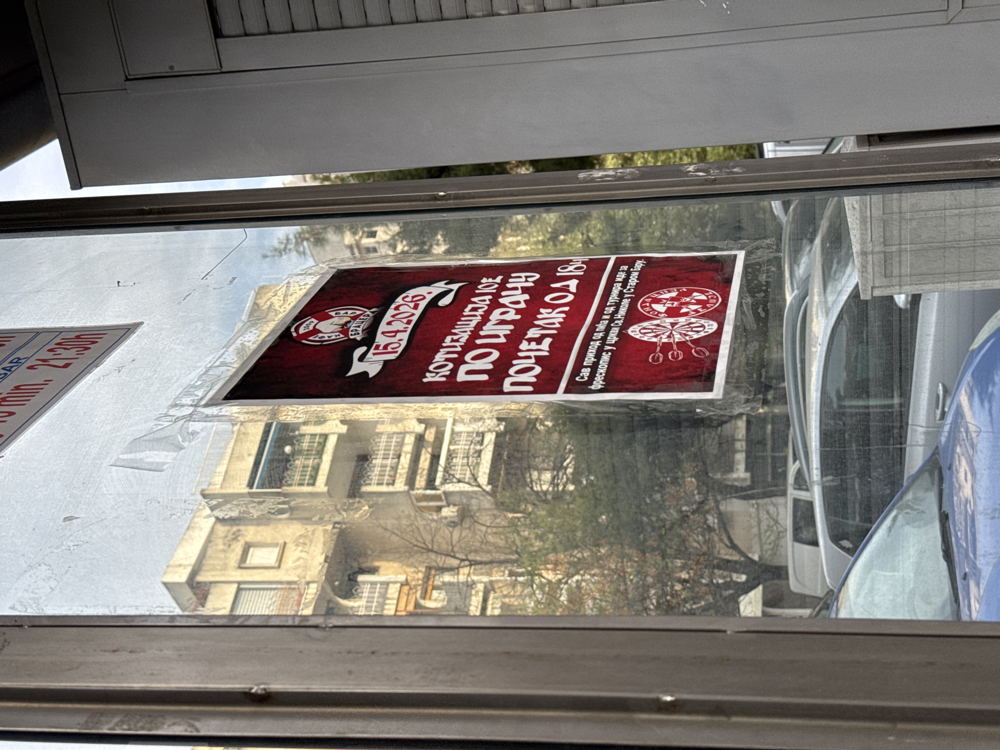 The image depicts a poster affixed to a window, likely a bus shelter. The poster advertises a dart tournament scheduled for April 15, 2026, with entry fees per player and a start time of 6:00 PM. The poster indicates that all proceeds from the sale of drinks and the tournament will be used to fund the restoration of frescoes in the Church of St. Nicholas in Stari Bar, Montenegro. A bus schedule is visible to the left of the tournament poster. The reflection in the window includes a view of a building with windows and a car parked nearby.