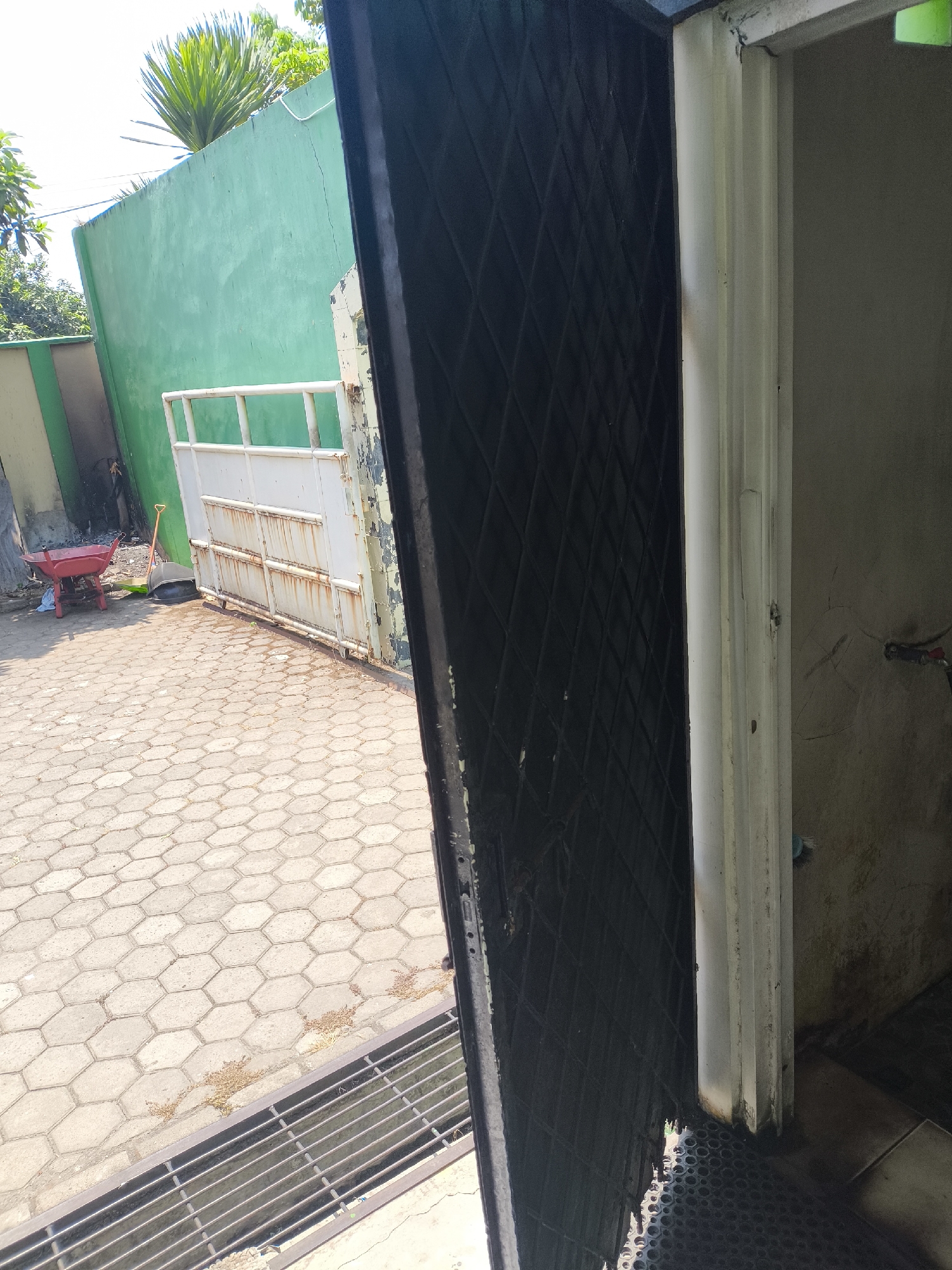 The image shows an open black metal security door in the foreground, angled to the right, revealing a partial view of a light-colored door frame and a plain wall inside. The exterior environment, visible to the left, features a hexagonal paver stone ground surface. A red wheelbarrow is present on the pavers, along with a broom and dustpan. Beyond the pavers, a white metal gate stands in front of a green wall. Trees are visible behind the green wall, and the sky is bright overhead, suggesting daytime. The scene appears to be an outdoor area, possibly a courtyard or pathway, adjacent to a building. Based on the distinctive green wall and the style of the paving and gate, this scene is consistent with outdoor common areas found in residential or commercial properties within Cilegon, Indonesia.