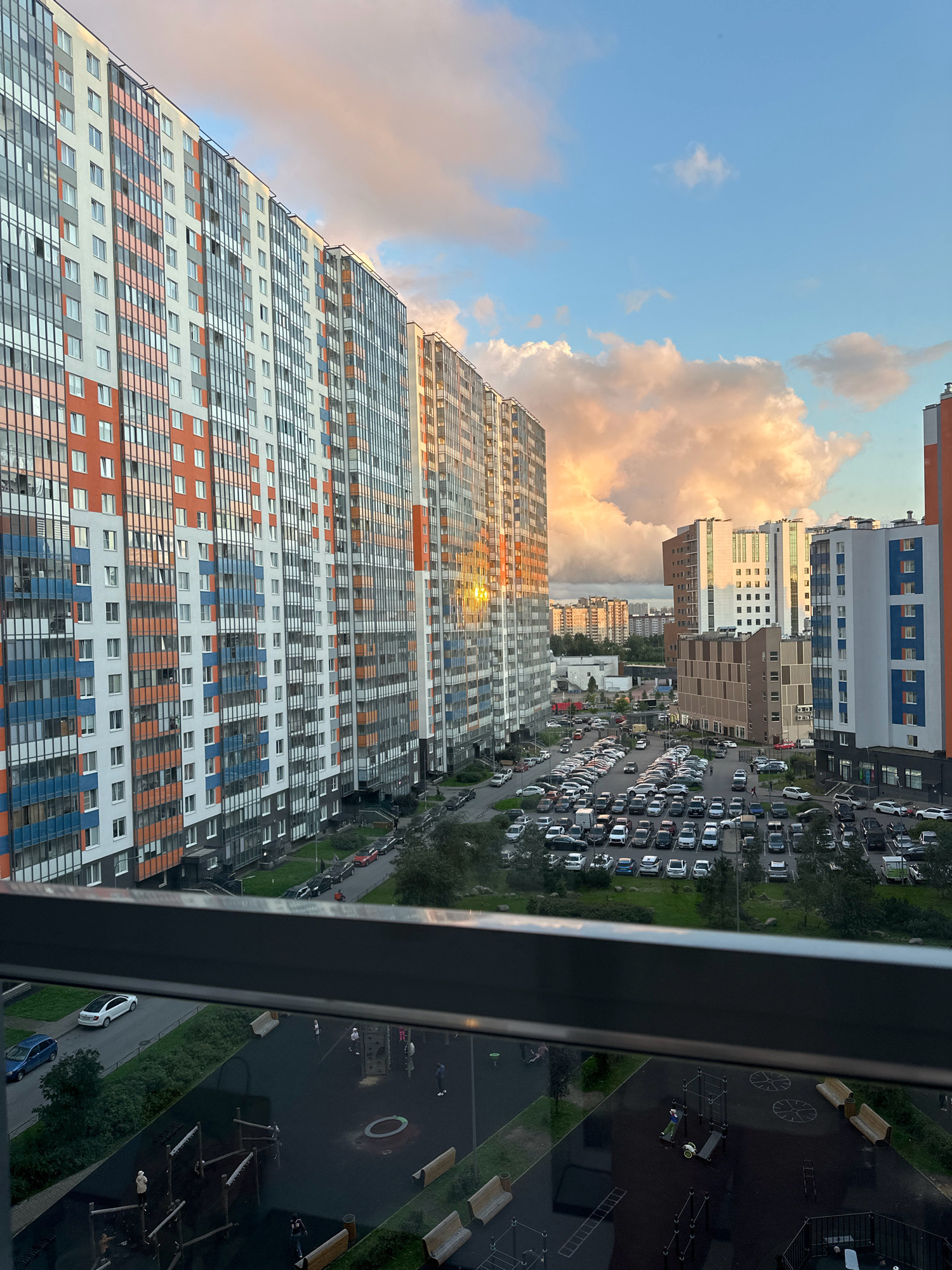 The image captures a street scene in a residential area of Saint Petersburg, Russia, likely during the golden hour due to the warm lighting on the buildings and clouds. Dominating the foreground are several multi-story apartment buildings with a modern architectural style, featuring balconies and a mix of white, orange, and blue facades. A large, packed parking lot filled with numerous cars is visible between the buildings. In the lower part of the image, a playground area with various play structures and benches is depicted. The sky is partly cloudy, with some clouds illuminated by the setting sun, creating a dramatic visual effect. A metal railing runs along the bottom edge of the frame, suggesting the photo was taken from a balcony or elevated viewpoint.