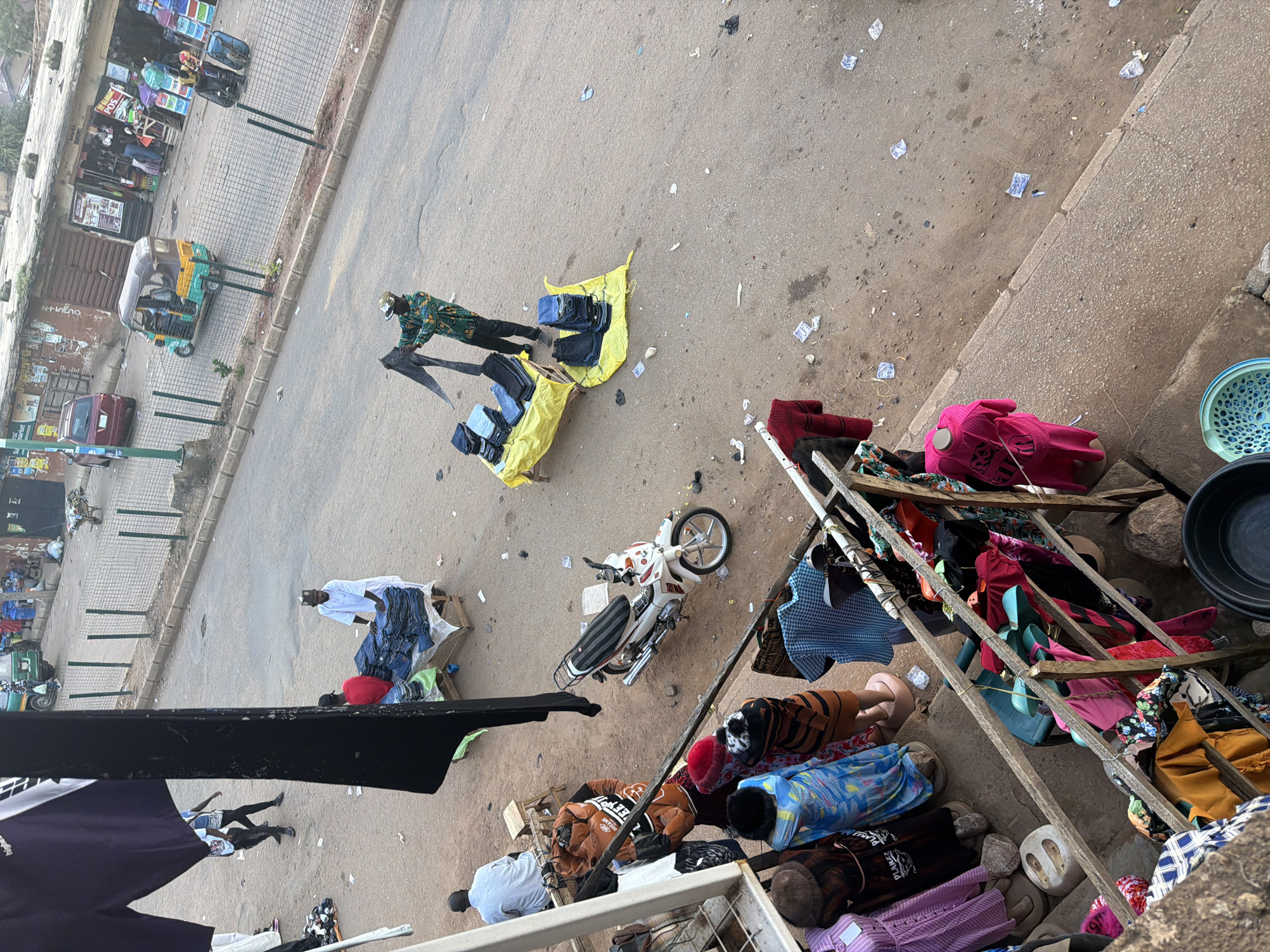 This image depicts a bustling street market scene in Bukuru, Nigeria. The foreground shows a stall with various clothing items displayed on mannequins and racks. Further back, a man in a white shirt and cap is seen arranging stacks of jeans. Across the street, a vendor in a patterned shirt is holding up a pair of jeans, seemingly showcasing them to potential buyers. Several other individuals are scattered throughout the scene, engaged in various activities. A three-wheeled vehicle, commonly known as a "keke," is visible in the middle ground, alongside parked motorcycles and a red car. The environment appears to be a typical urban street with shops lining one side and a sidewalk, all set against a backdrop of buildings. The overall impression is one of vibrant commerce and daily life unfolding in the Nigerian city.