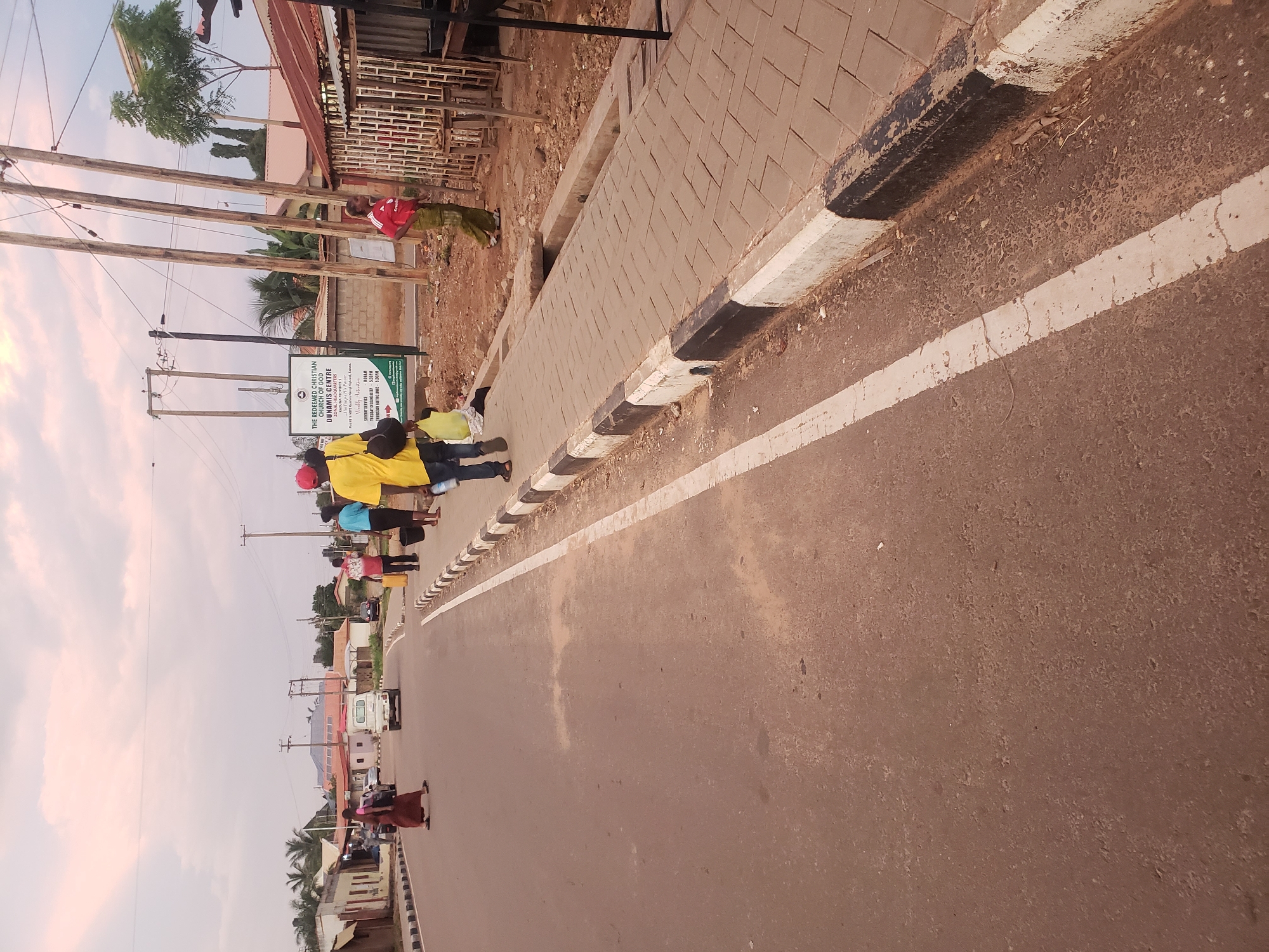 The image shows a street scene in Narayi, Nigeria, likely during the late afternoon or early evening, given the lighting. The road is asphalt with a white line marking the lane. A paved sidewalk runs alongside the road, bordered by a curb painted with alternating black and white stripes. Several people are present. On the road, a person in a red dress is walking. On the sidewalk, a person in a yellow shirt and red cap is walking away from the camera, carrying a black bag and a yellow bag. Another person in a blue shirt and black pants is walking next to them, and further down the sidewalk, a person in a red shirt and green pants is standing. A white utility vehicle is visible on the road. In the background, there are buildings, utility poles, and trees. A sign for "THE REDEEMED CHRISTIAN CHURCH OF GOD DUNAMIS CENTRE" is also visible. The overall environment appears to be a moderately urban setting with some commercial activity.