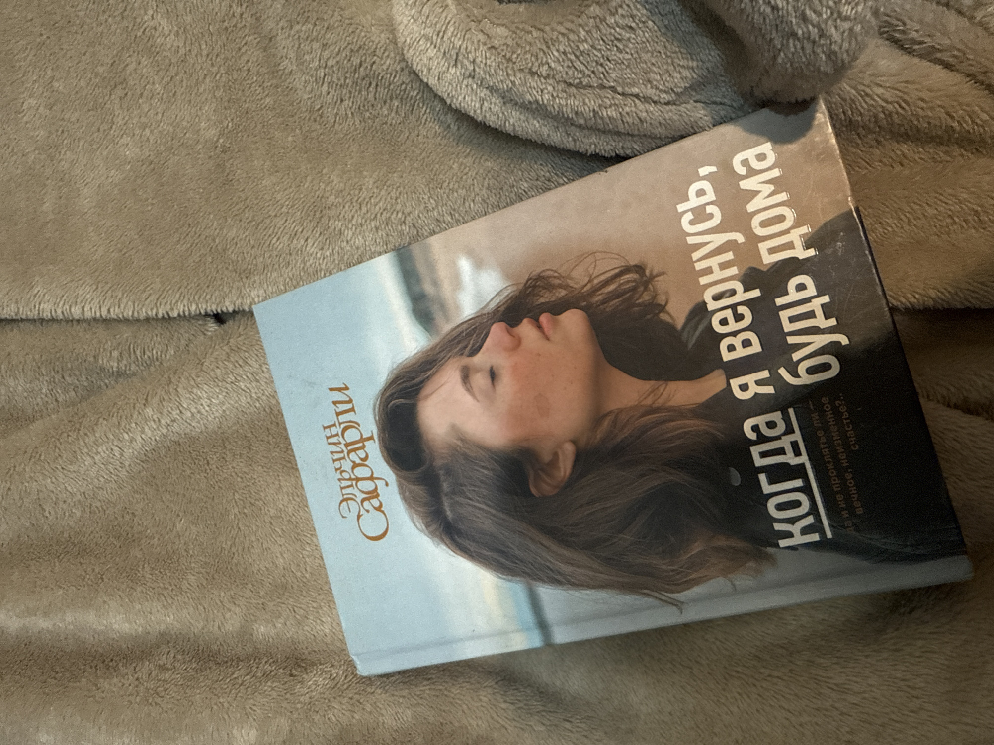 A book lies on a tan, plush blanket. The book's cover is light blue with a picture of a woman's face in profile. The title is in Russian: "Когда я вернусь, будь дома" (When I return, be home) by Elchin Safarli. The location is not discernible in the image.
