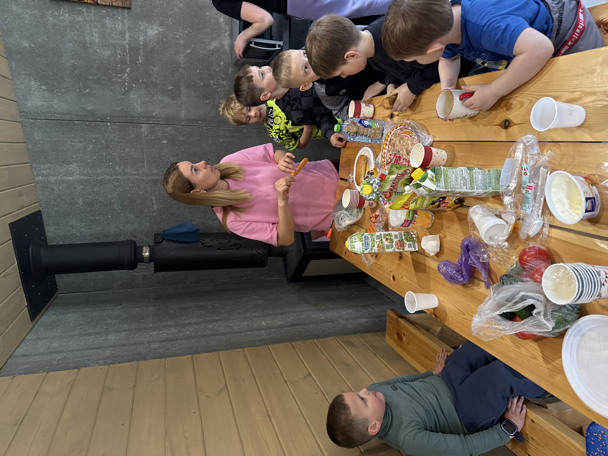 The image depicts an indoor gathering centered around a rustic wooden table, featuring an adult woman and five young boys. The woman, with long, light brown hair, wears a pink long-sleeved shirt and holds a sausage, seemingly demonstrating or offering it. The boys, ranging from young children to pre-teens, appear attentive. One boy in a blue shirt and grey shorts looks down at the table, another in a black long-sleeved shirt holds a ceramic mug, and two others in black shirts (one with a yellow pattern) watch the woman. A fifth boy, dressed in a grey turtleneck and dark pants, lies on a wooden bench beneath the table, observing the scene. Part of another adult is visible in the upper left corner.
The table is filled with various food and drink items. These include plastic bottles of water, several juice cartons (with Russian text, one mentioning "4 fruit"), plastic and patterned paper cups, bags of snacks (one clearly labelled "Карамельки" - Karamels/Candies), and plastic bags containing fresh vegetables like tomatoes and cucumbers. There's also a container of a white creamy substance and disposable cutlery. One white plastic cup has "МИР" (MIR - Peace/World) visible on its side. A boy's grey shorts have "KELY" on the waistband.
The room features a textured grey wall and light wooden plank flooring and wall panels, suggesting a simple or communal space. A black metal stove pipe or chimney extends from the wall near the woman. The activity appears to be a communal meal or snack time, with the children engaged in the process, possibly in a camp, community center, or casual home setting in Yekaterinburg, Russia. The children's expressions are generally focused and calm.