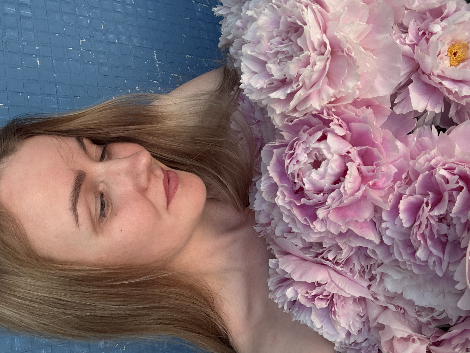 A close-up, head-and-shoulders portrait shows a fair-skinned woman with blonde hair and a light complexion. She is looking down and to her right, with her lips slightly parted. Her shoulders are bare and she is adorned with a large bouquet of pink peonies. The background is a textured blue wall. The image does not contain any information that would suggest a specific location in Chaikovsky, Russia.