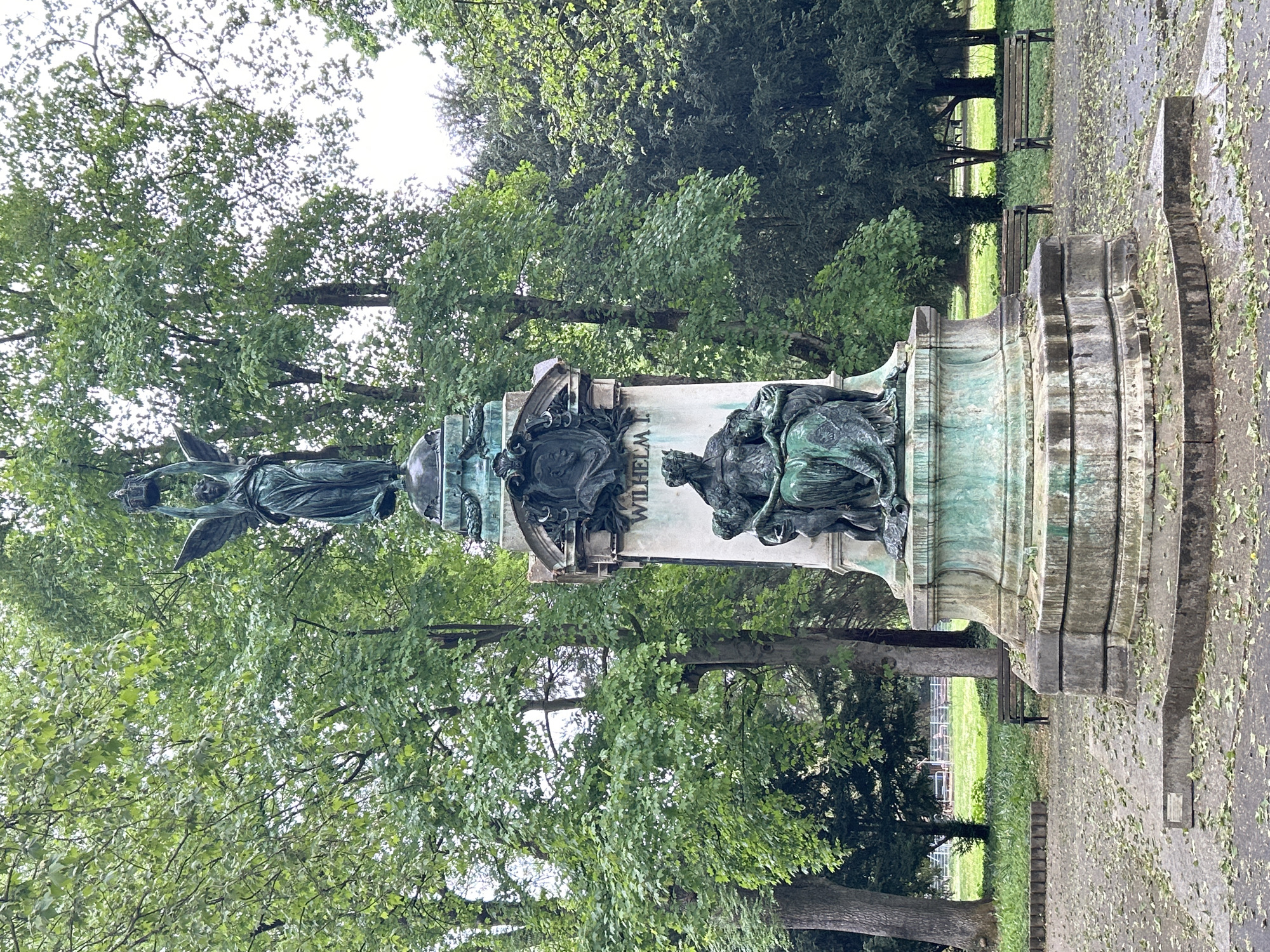 The image shows the Wilhelm I memorial in the Hofgarten park in Düsseldorf, Germany. The memorial is a large, ornate structure made of stone and bronze. It features a statue of Wilhelm I on top, surrounded by other figures and decorative elements. The memorial is located in a park-like setting, with trees and benches visible in the background.