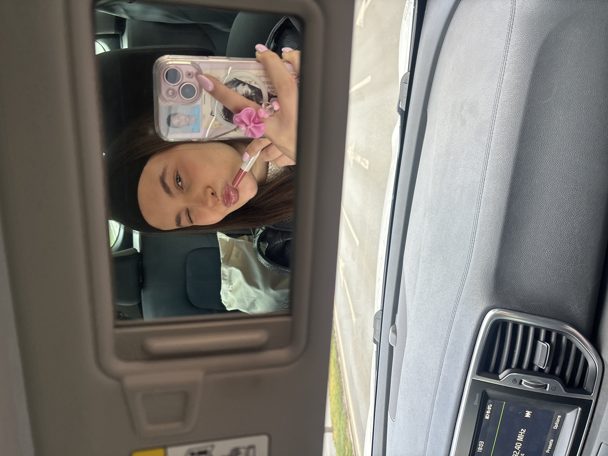 A woman is taking a selfie in a car mirror. She is wearing a black headband and putting on lip gloss. Her phone has a clear case with a photo of a person and a pink flower ring attached. The car's dashboard is visible, showing the radio is tuned to 92.40 MHz, which is the frequency for "BG Radio" in Sofia, Bulgaria. The time on the display is 18:03. Through the car window, a street can be seen.