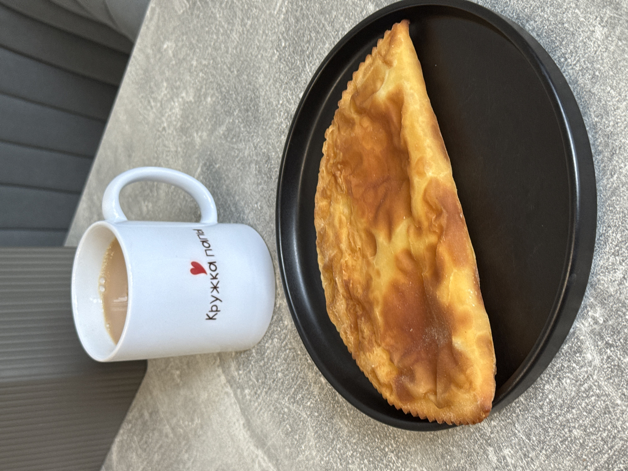A white mug with a red heart and Cyrillic text that reads "Кружка папы" (Dad's mug) sits on a textured gray surface next to a half-eaten cheburek on a black plate. The cheburek is a golden-brown, fried pastry with a crimped edge, its filling partially visible. The mug contains a light brown liquid, likely tea or coffee, with some bubbles on the surface. The background shows a blurry gray ribbed surface, possibly a chair or furniture. The scene is likely indoors, on a table, in a cafe or home environment, potentially located in Lyubertsy, Russia, given the text on the mug.