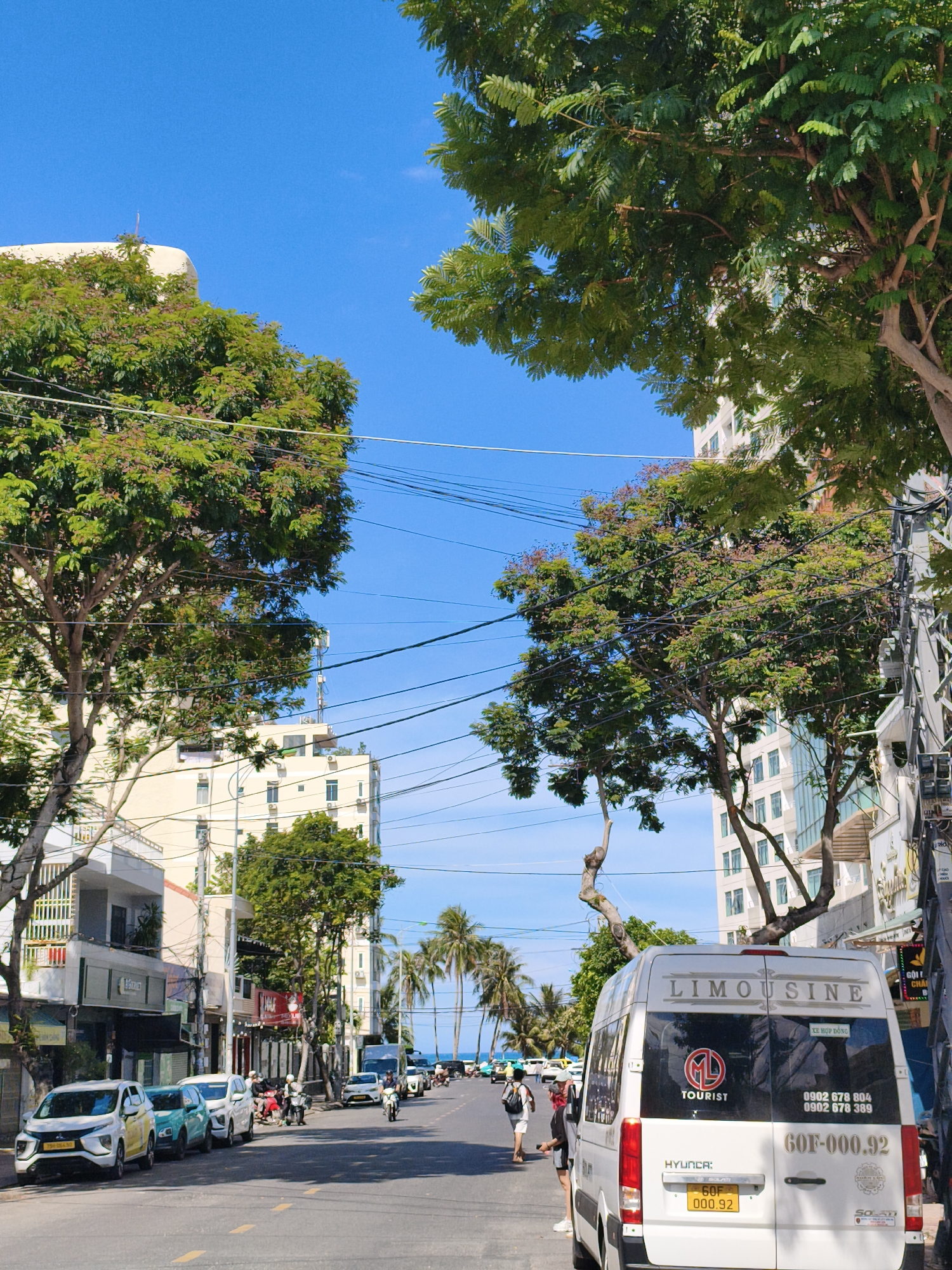 In Nha Trang, Vietnam, a street scene unfolds under a clear blue sky, bordered by lush green trees and multi-story buildings. A white van, identified as a "LIMOUSINE" and "TOURIST" vehicle, is parked on the right side of the road. Its license plate reads "60F-000.92". Two individuals, one wearing a red bandana, stand near the van. The street is lined with parked cars and motorcycles, and palm trees are visible in the distance, suggesting proximity to a coastal area. Buildings on either side of the street display various signs and advertisements. The road surface is asphalt with yellow lane markings.