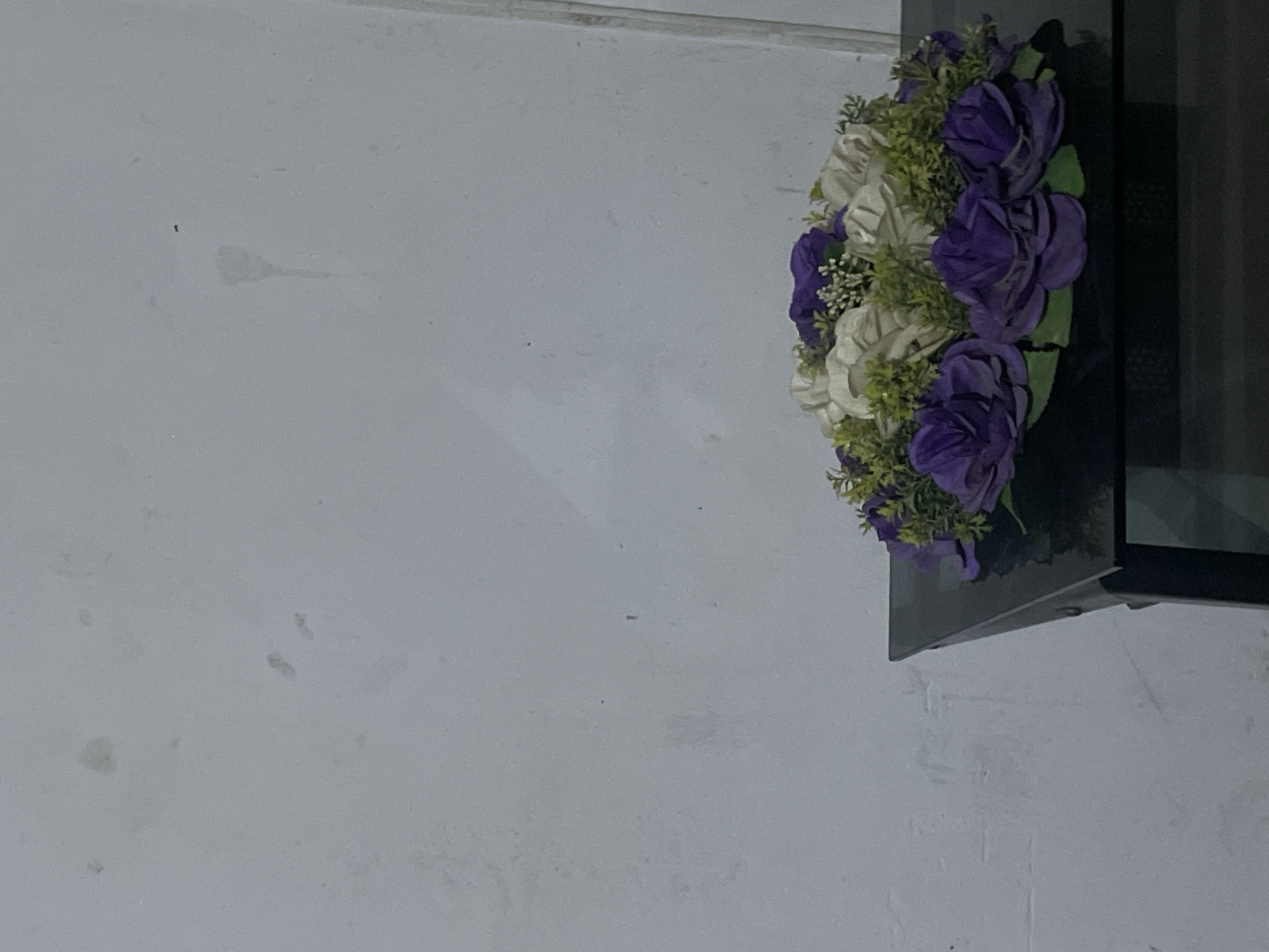 A cluster of artificial white and purple flowers with green foliage is positioned on a gray wall next to a black, framed opening. The location, Jos, Nigeria is not determinable from the image.