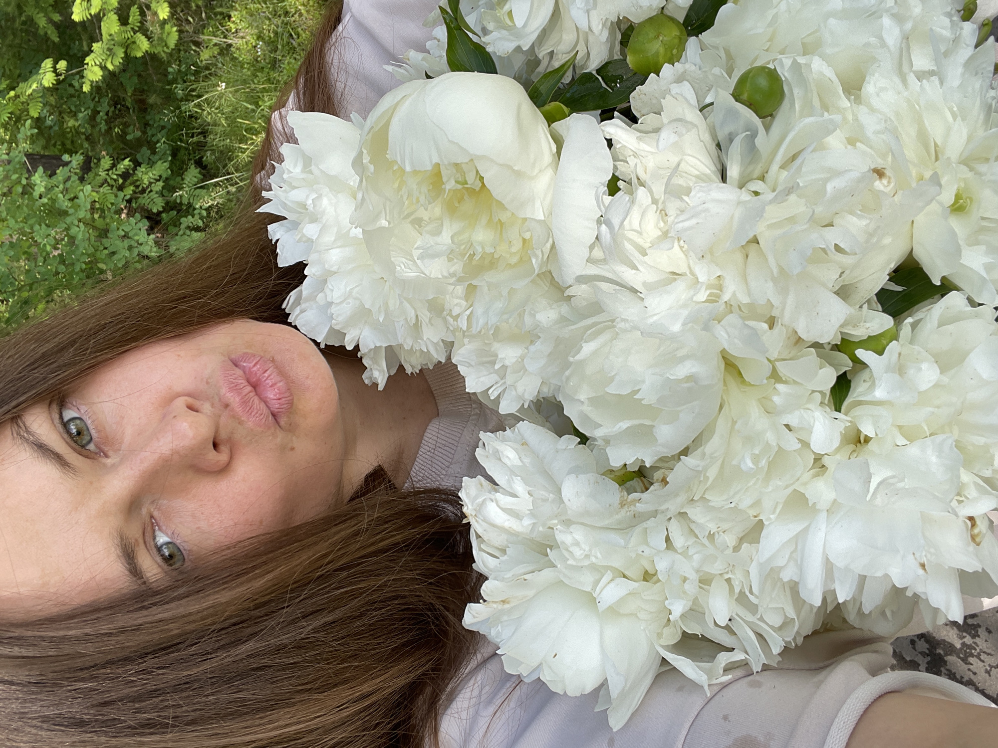 A woman with long brown hair and green eyes is holding a bouquet of white peonies. She is wearing a light-colored sweatshirt. The background consists of lush green foliage, suggesting an outdoor, natural setting, possibly a park or garden. The image captures a close-up view of the woman and the flowers, with the background slightly blurred. The woman is puckering her lips, creating a playful or affectionate expression. The overall impression is one of natural beauty and softness. The city of Заречный, Russia, is mentioned as the location.