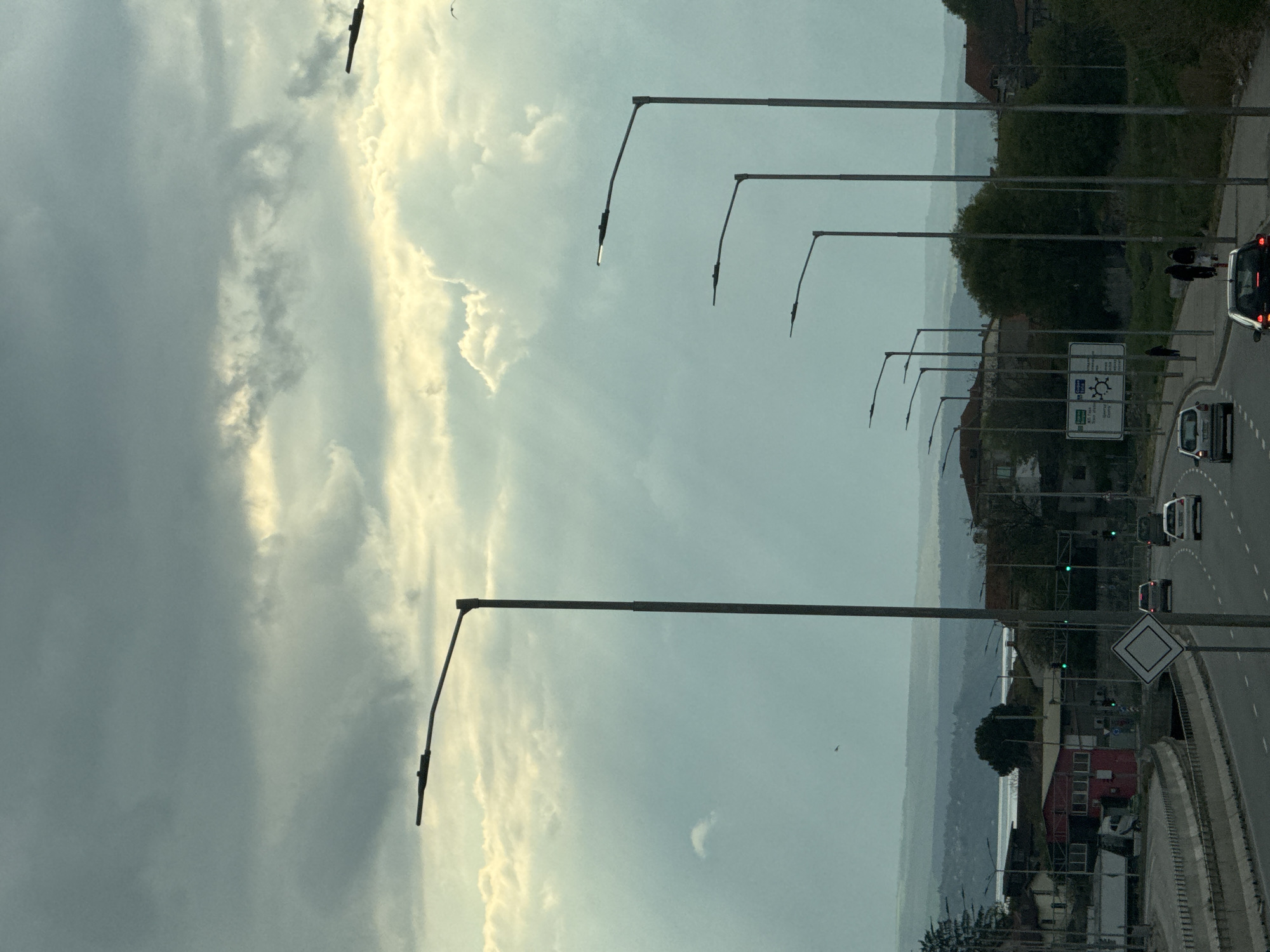 This image, taken in Varna, Bulgaria, depicts a road scene under an overcast sky. The photograph is vertically oriented but shows a multi-lane road or highway curving from the lower-left towards the upper-right. A prominent row of modern streetlights extends along the right side of the road, with some individual lights appearing very faintly illuminated or reflecting light.
Several vehicles are on the road, including a white van in the foreground, a dark car with illuminated brake lights further up, and another white car in the distance. Two pedestrians are visible walking on what appears to be a sidewalk or shoulder adjacent to the road. Beyond the road on the right, residential buildings, including a distinctive red-roofed structure, are interspersed with trees and bushes. In the hazy distance, a large body of water, likely the Black Sea, meets distant hills or landmasses.
The sky is largely covered with clouds, though patches of brighter, yellowish light break through, suggesting either late afternoon or early morning, or simply reflections on an overcast day. A few small birds are seen flying.
Notable road signs include a large white directional sign with "E87", "Варна" (Varna), and "Шумен" (Shumen) clearly visible, along with a blue circular symbol indicating a roundabout. A white diamond-shaped sign, denoting a priority road, is also present. The scene captures an everyday urban-coastal environment with regular road traffic and pedestrian activity.