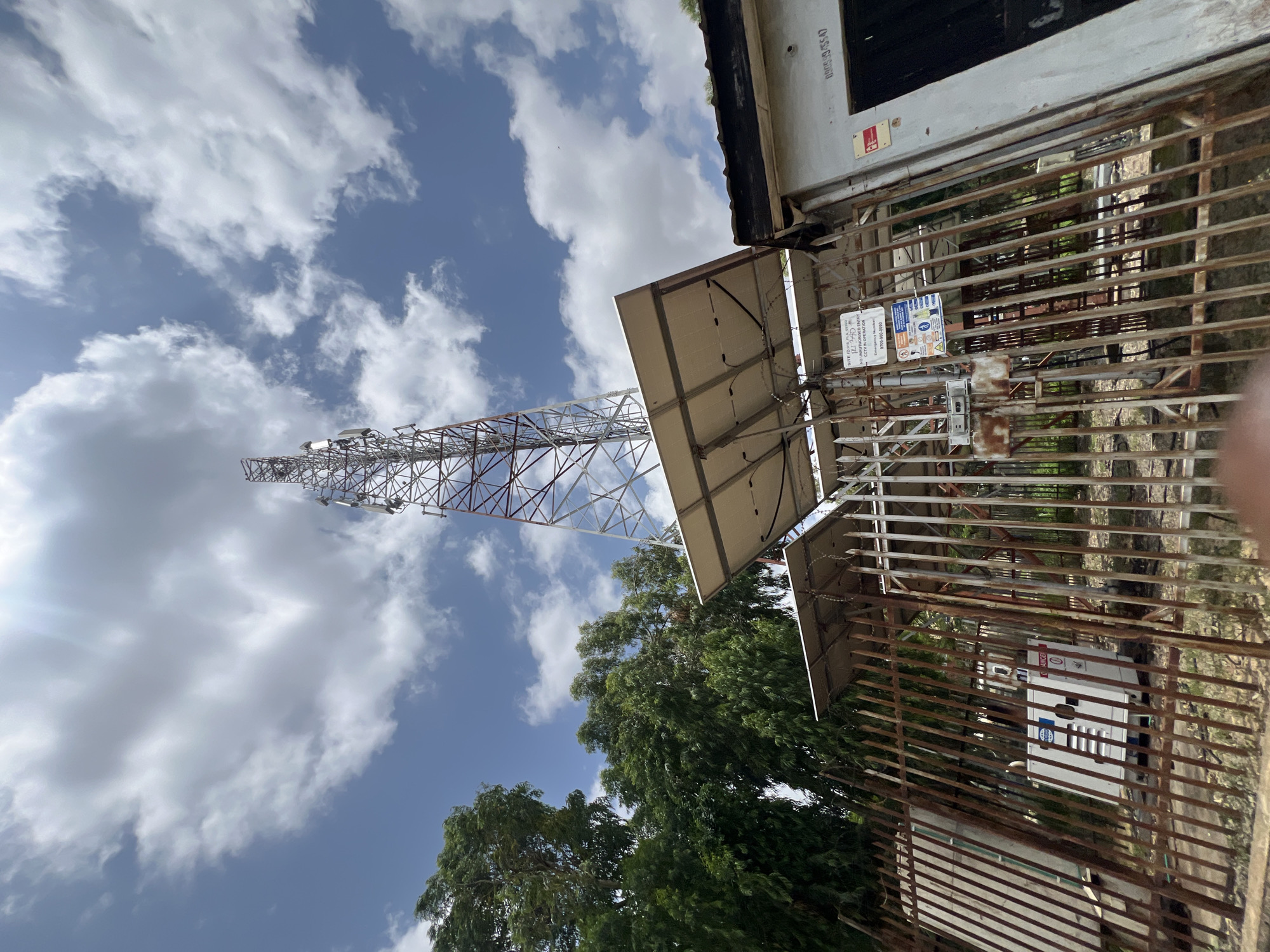 This is a low-angle, full outdoor shot of a telecommunications tower in Jos, Nigeria. The tower is a tall, lattice structure with several antennas mounted at different levels. It is surrounded by a rusty metal fence. In the foreground, there are several solar panels mounted on metal frames, likely to power the tower. Behind the fence, there is a white generator unit. To the left of the tower, there are dense green trees, and to the right, a small building with a dark door and a sign. The sky is blue with scattered white clouds, and the sun is shining brightly, casting shadows. The overall scene depicts an operational telecommunications site.