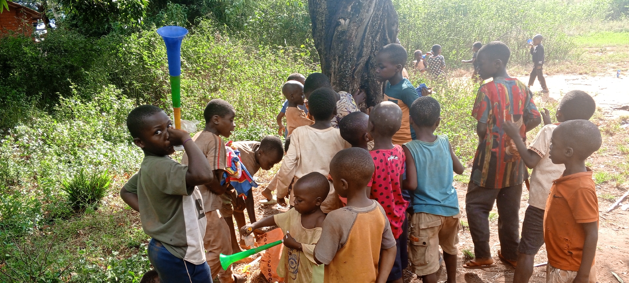 A group of children stand in a field in Ngalula, Democratic Republic of the Congo. Some children are looking at the ground, others are looking at the camera. One child is holding a tall, multi-colored plastic trumpet-like object, and another holds a green watering can. A large tree is behind the group, and foliage is visible in the background. One child in the background is walking away.