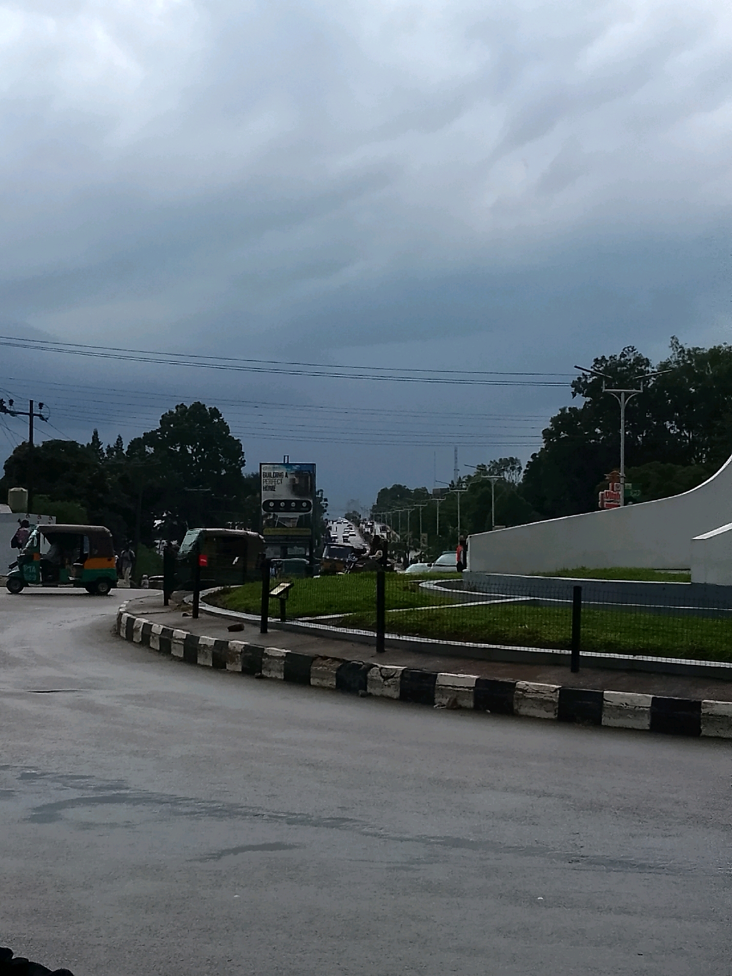 The image depicts a cloudy day scene in Jos, Nigeria, likely at a roundabout. In the foreground, a paved road curves to the left, with a black and white striped kerb marking the edge. To the left of the road, a three-wheeled auto-rickshaw, often called an "okada" or "keke," is parked. A person can be seen in the driver's seat. Further to the left, behind the auto-rickshaw, is a white building with a black roof.
In the middle ground, a traffic light is visible, showing a red signal. Beyond the traffic light, the road continues, with several vehicles, including cars and trucks, visible in the distance, suggesting moderate traffic. A billboard with an advertisement for "BUILDING A PERFECT HOME" is positioned on the right side of the road. Green grass and some trees are visible behind the kerb and surrounding the roundabout area. In the background, more trees and power lines are present under a grey, overcast sky. The overall impression is of a typical urban street scene in Nigeria on a cloudy day.