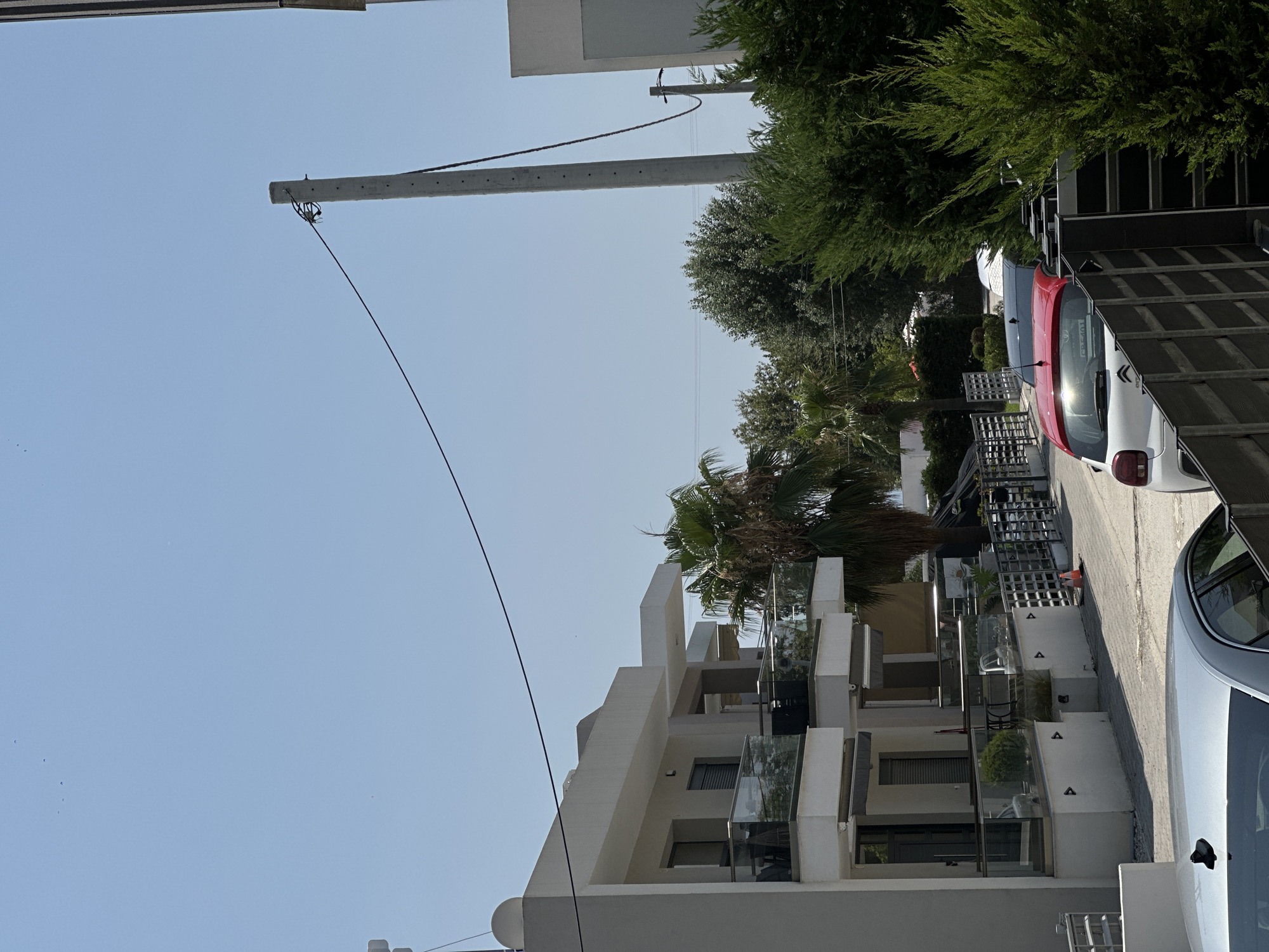 A street scene in Ofrynio Beach, Greece, shows a multi-story building with balconies and awnings on the left, and dense green trees and foliage on the right. A concrete utility pole stands tall in the center, with a wire extending from it across the sky. Several cars are parked along the street, including a red and white car, and a silver car partially visible in the foreground. The sky is clear and blue.