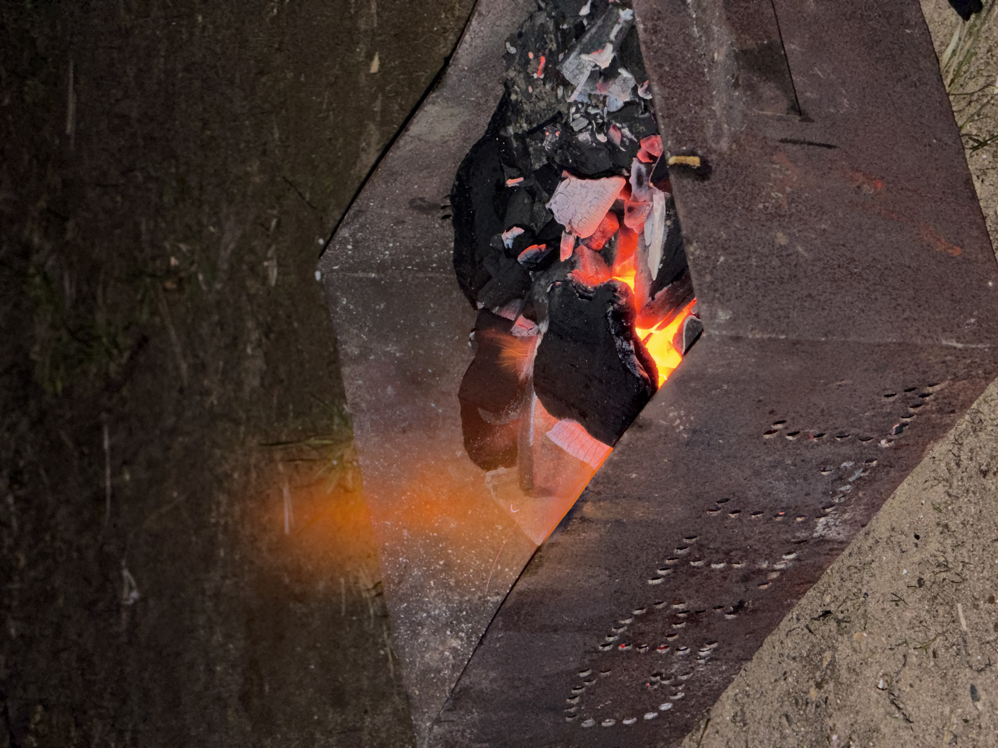 The image captures a close-up view of a metal brazier or mangal containing burning charcoal and glowing embers, suggesting an outdoor setting at night or in very low light. The central feature is the intensely bright, reddish-orange and yellow glow of the embers and charcoal, which are nestled within a dark, angular metal structure. The burning fuel provides the primary illumination for the scene.
The metal structure itself appears to be made of sturdy, possibly rusted, sheet metal. On its right side, numerous small, crescent-shaped perforations are visible, arranged in a patterned design. These holes likely serve as ventilation for the fire, and their edges are faintly illuminated by the internal glow. The left side of the metal structure is also dark and angular, reflecting some of the fire's warmth.
The ground surrounding the brazier is dark and indistinct due to the low light conditions. On the left side, hints of dark grass or foliage can be seen, partially illuminated by a soft, orange glow cast by the embers. On the right, the ground appears to be dark earth or sand. The overall impression is one of a rustic, outdoor environment, possibly preparing for grilling or providing warmth, in Nikolo-Beryozovka, Russia. There is no legible text visible in the image.