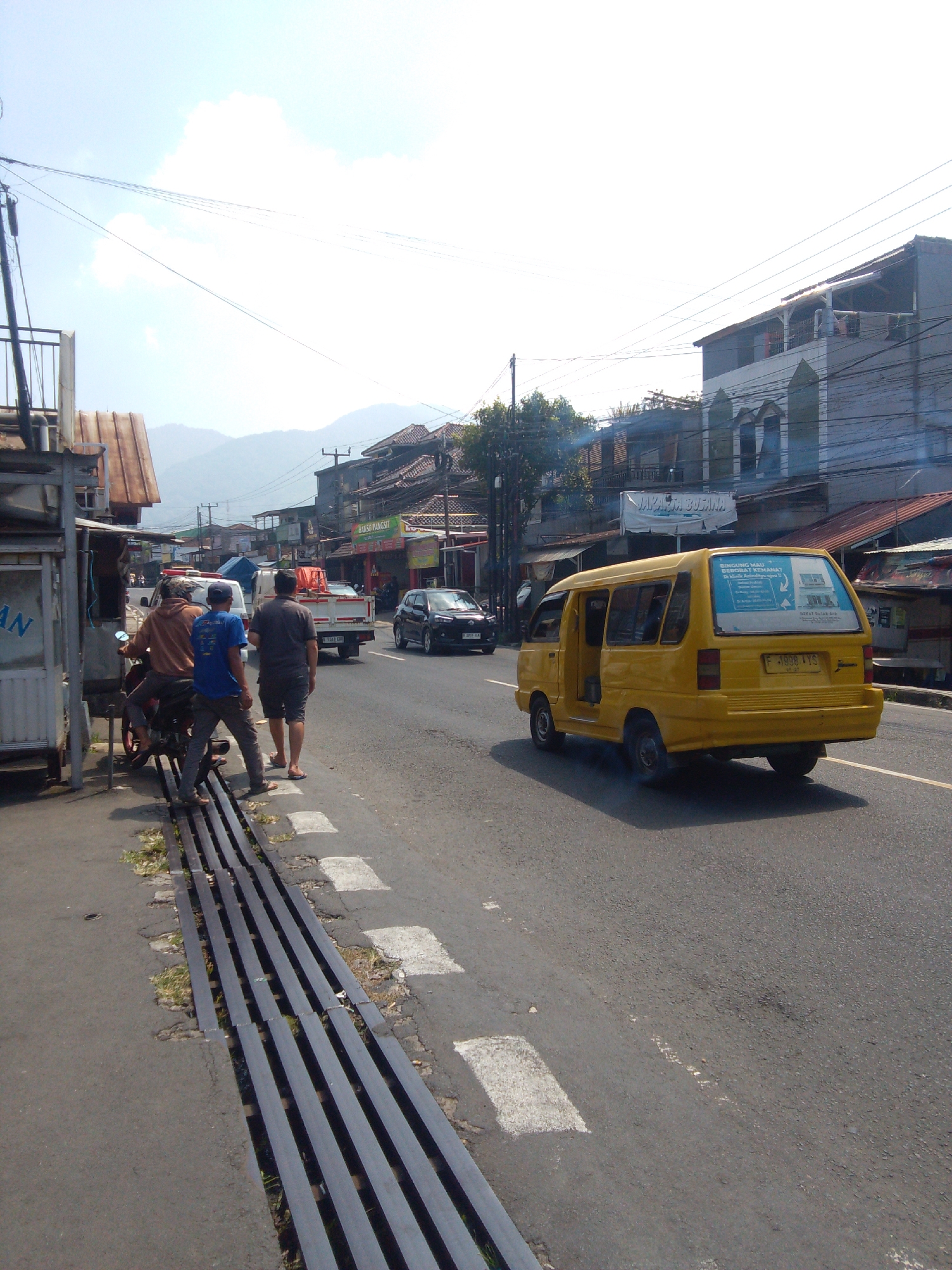 The image depicts a street scene in Cianjur, Indonesia. In the foreground, three men are standing near a motorcycle. One man, wearing a brown hoodie and a cap, is seated on the motorcycle. Another man, in a blue t-shirt and grey pants, is standing next to the motorcycle, and a third man, wearing a dark grey t-shirt and dark shorts, is standing further to the right.
The street is paved and has a metal grate running along the side. In the background, there is a yellow van driving on the road, with Indonesian license plates. A black car is also visible further down the road, and a white truck is parked. Buildings line the street, some appearing to be shops or businesses, with signs in Indonesian visible. Hills or mountains are visible in the distance. The sky is bright and appears to be a sunny day.