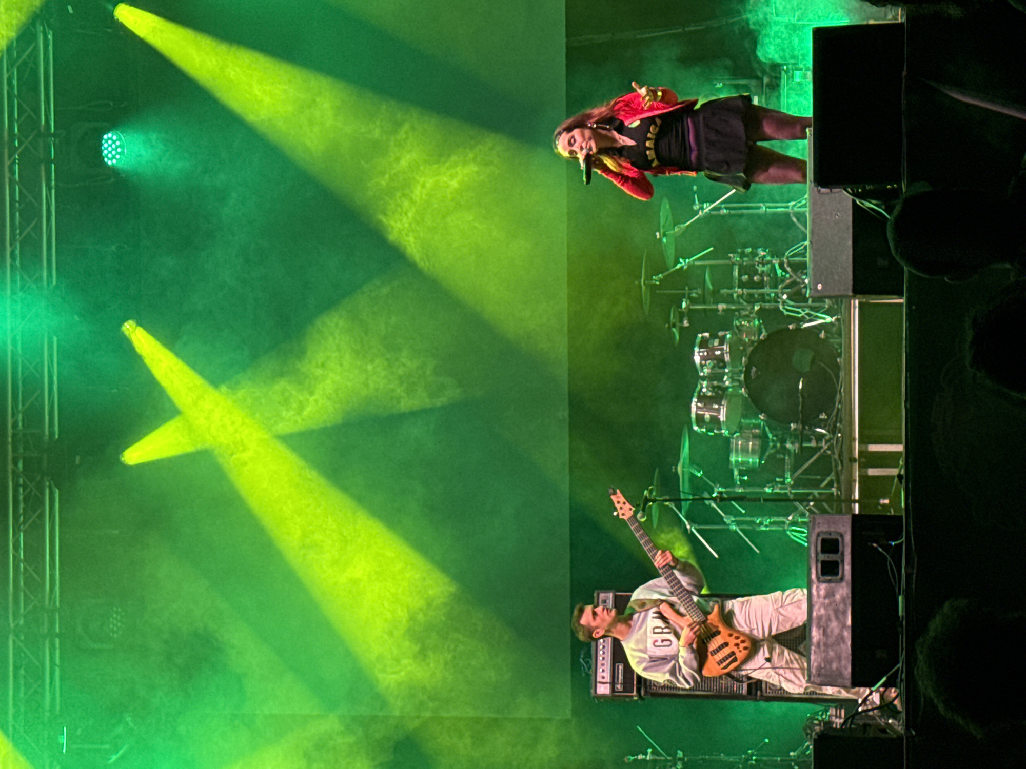The image shows a live music performance, likely at the Vida Festival in Vilanova i la Geltrú, Spain. A female vocalist in a red jacket and black dress is singing into a microphone. A male bassist in a white sweater and pants is playing his instrument. A drum set is visible in the background. Green stage lights illuminate the scene. Loudspeakers are also visible.