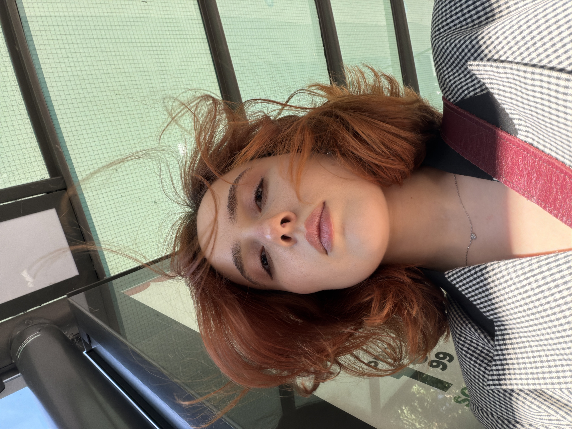 A young woman with short, wind-swept auburn hair is shown from a low angle, looking up at the camera. She is wearing a black and white checked blazer and a delicate silver necklace with a small heart pendant. Her skin is fair, and she has subtle makeup on. The background consists of a light green, gridded ceiling and a metal pole, suggesting she is at a bus stop or shelter. Reflections of trees and possibly buildings are visible on the green ceiling. There are no other people in the frame. The scene is outdoors, likely during the daytime, given the bright lighting and shadows. The specific location in Strasbourg, France, cannot be determined from the visual information provided.