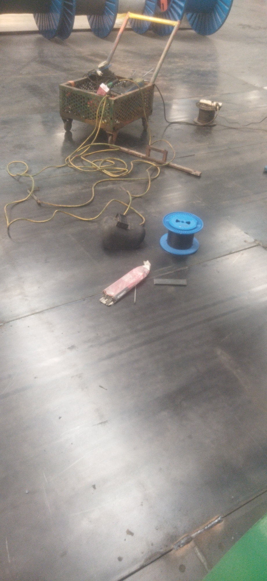 The image depicts a cluttered indoor scene, likely a workshop or industrial setting. In the foreground, a dark gray floor with visible seams runs across the frame. On the floor, a blue spool of wire sits next to a dark, amorphous object with a yellow wire extending from it. Further to the left, a pink and red package is lying on its side, with a thin wire or rod protruding from it. A thick yellow cable snakes across the floor from a green, mesh-like cart. This cart, supported by four legs, contains various items, including what appear to be electrical components and more wiring. In the background, large, blue cylindrical objects, possibly industrial spools or machinery, are partially visible. The lighting is subdued, suggesting an indoor environment without direct sunlight. No individuals are present in the image. The overall impression is one of a work-in-progress or a storage area containing materials and equipment for electrical or mechanical tasks.
