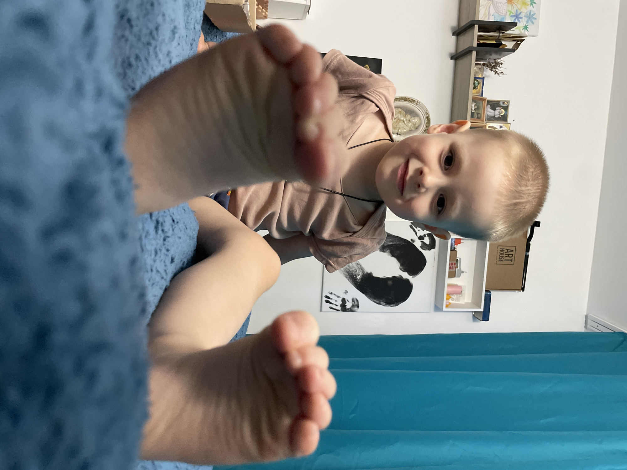 A boy is angled in the center of the image, peering down with a smile. He has short, blonde hair and is wearing a light brown shirt with a thin necklace. The foreground is dominated by a person's feet, slightly out of focus, resting on a textured blue blanket. Behind the boy, there is a white wall with a small shelf containing various objects and framed pictures. A white canvas with black hand and footprints can also be seen. To the right of the boy, there is a turquoise curtain. The location is Unknown, Unknown.