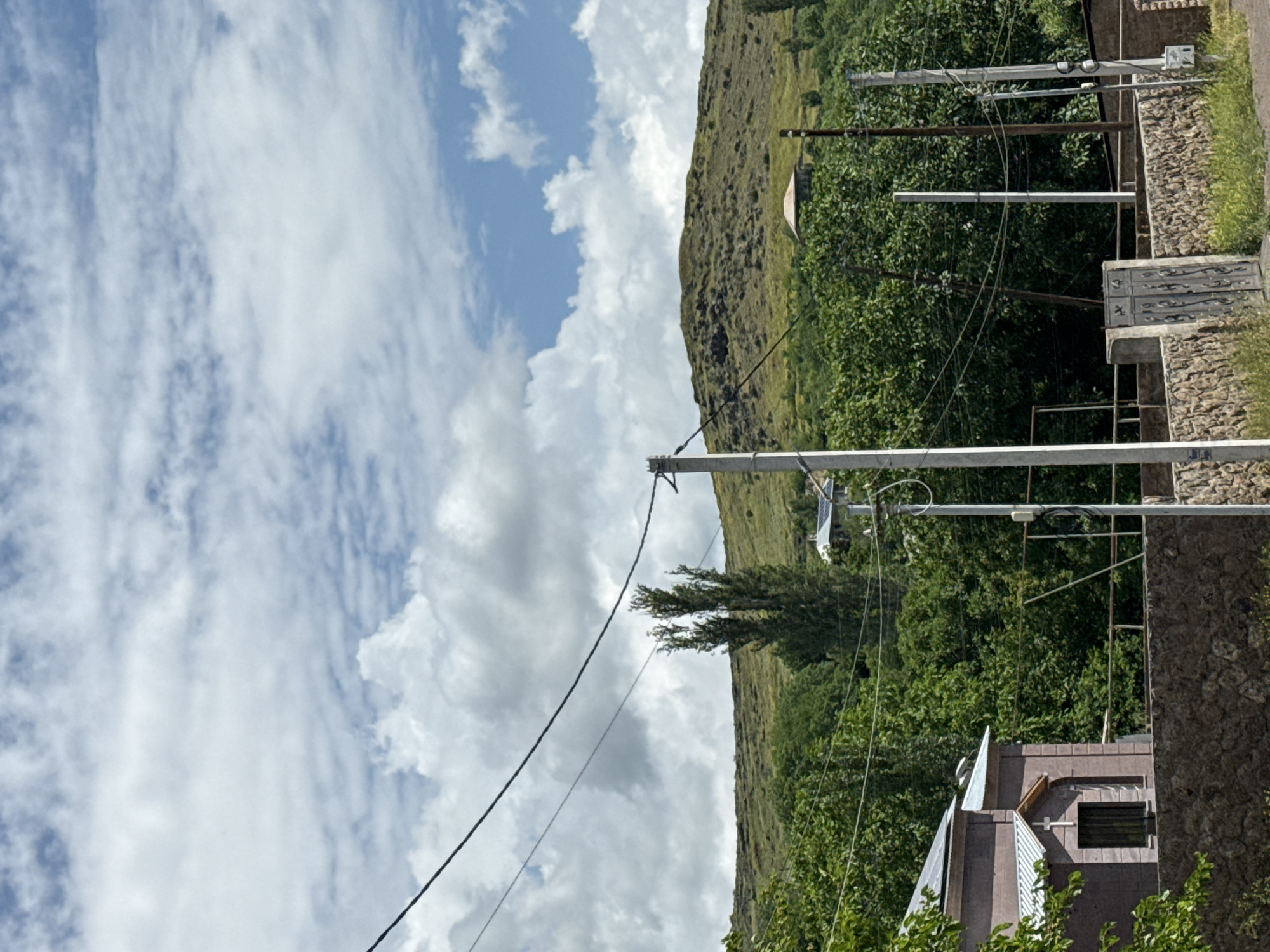 The image depicts a rural landscape under a cloudy sky. In the foreground, a stone wall runs horizontally across the frame, with a grey metal gate visible on the right side. To the left of the gate, a portion of a building with a light brown facade and a metal roof is visible. A small cross is mounted above a dark window on this building. Power lines traverse the scene from the top left, supported by utility poles.
Behind the wall and building, a dense line of green trees forms a backdrop. Further in the distance, a rolling hill covered in dry grass and scattered dark rocks rises up, its summit partially obscured by the clouds. A small, circular roof structure is visible on the slope of this hill. Two tall, slender trees stand out against the hill in the middle ground. The overall impression is one of a quiet, possibly isolated, rural setting.