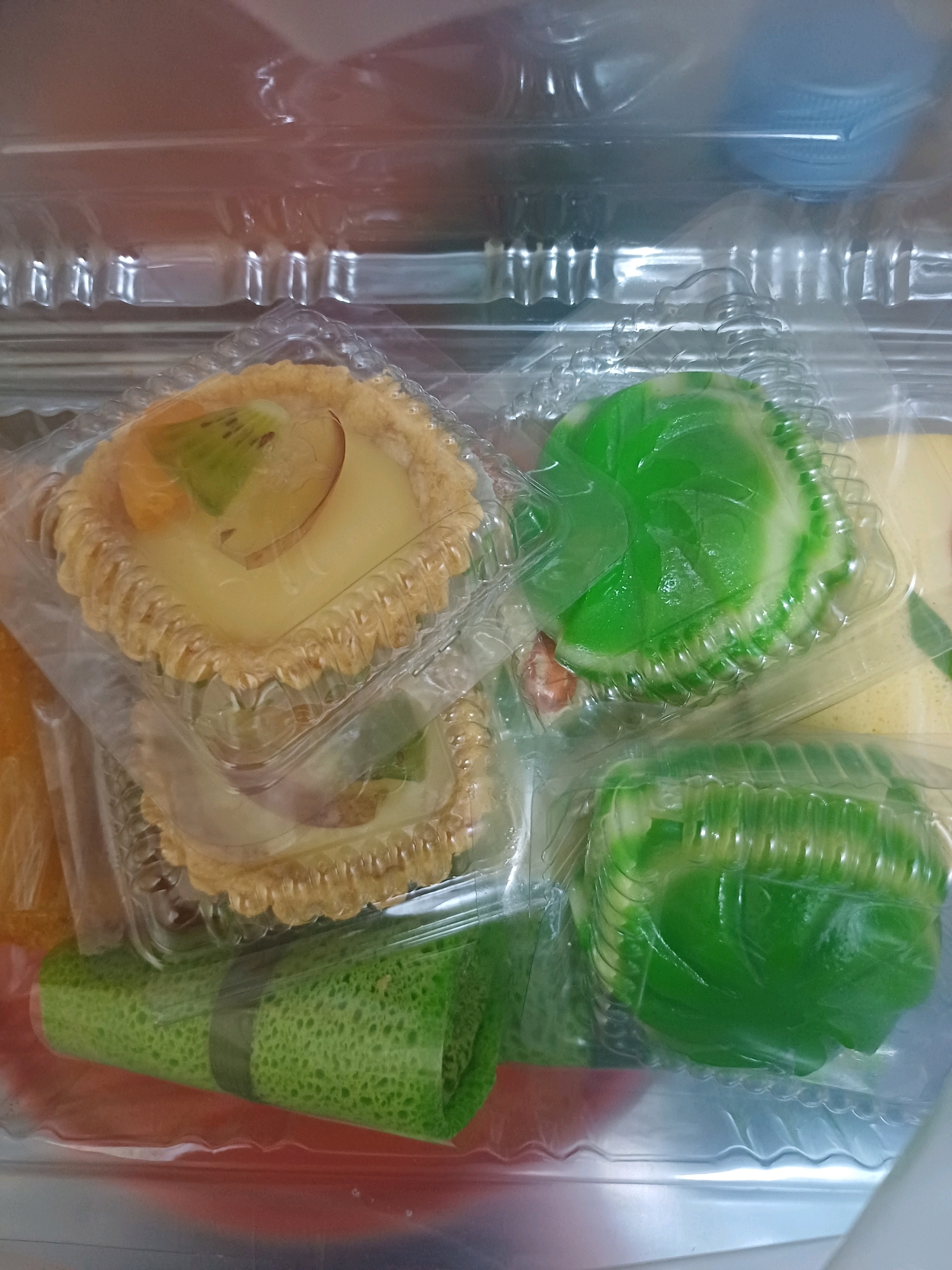 In Dumai City, Indonesia, this image captures a close-up view of various pastries and desserts packed in clear plastic containers. The items appear to be arranged in layers within a larger plastic casing.
In the foreground, a green rolled dessert with a textured surface is visible, secured with a dark band. Behind it, a round green jelly dessert with a marbled pattern is nestled in a fluted plastic container. To the left and slightly above, a fruit tartlet filled with a creamy custard and topped with slices of kiwi fruit and possibly apple is presented in a similar fluted container, stacked atop another tartlet of the same kind. The tartlets have a golden-brown, crimped crust.
The overall environment suggests a retail or home setting where these treats are packaged for sale or consumption. The lighting is soft, and the focus is on the food items, with the background slightly blurred, hinting at the transparency of the plastic packaging.