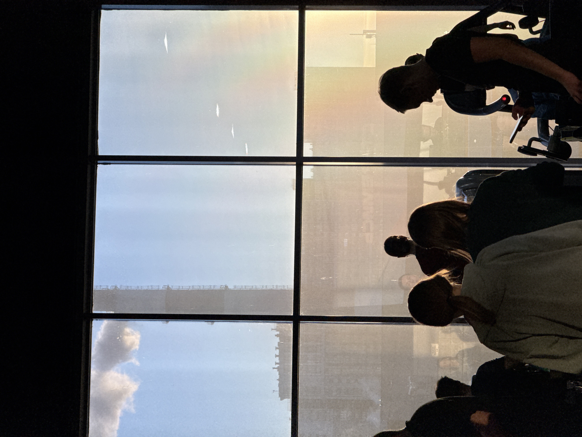 The image shows a view from a high-rise window. The window is divided into panes. The sky is visible through the window, with some clouds. Reflections of people are visible in the window panes. One person is sitting in an office chair and using a tablet. The general setting is an office or commercial space in a skyscraper. The presence of distinct architecture in the background, coupled with light, suggests it is Saint Petersburg, Russia.