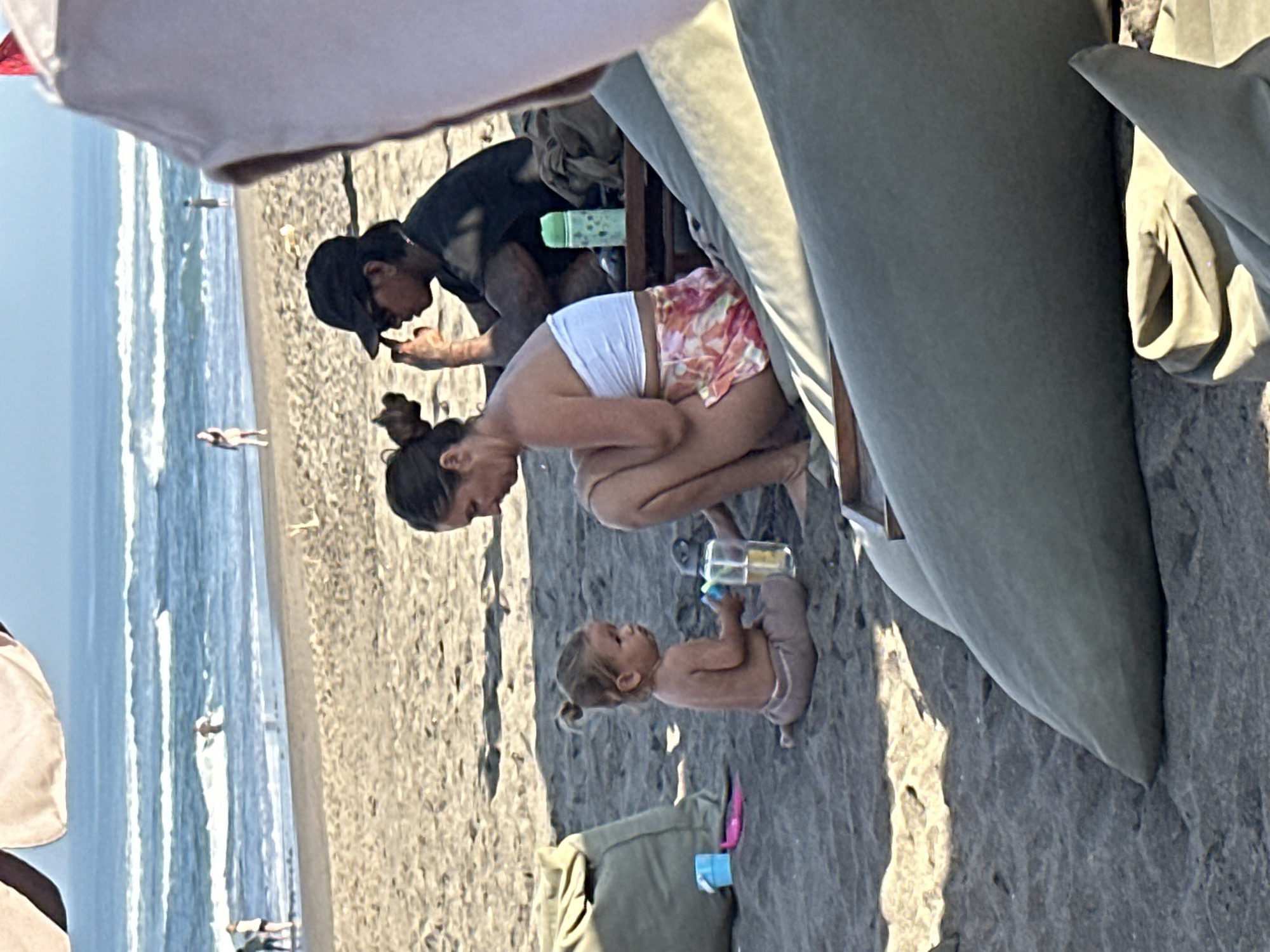 The image depicts a beach scene in North Kuta, Indonesia, with several people and objects on the sand. In the foreground, a woman in a white strapless top and patterned shorts sits on the sand, facing a young child. The child, with their hair in a bun, is also sitting on the sand, holding a toy and a cup. Behind them, another person wearing a black t-shirt and a baseball cap is seated. In the background, the ocean with waves is visible, and a few people can be seen walking on the shoreline. Beach chairs and bags are also present on the sand.