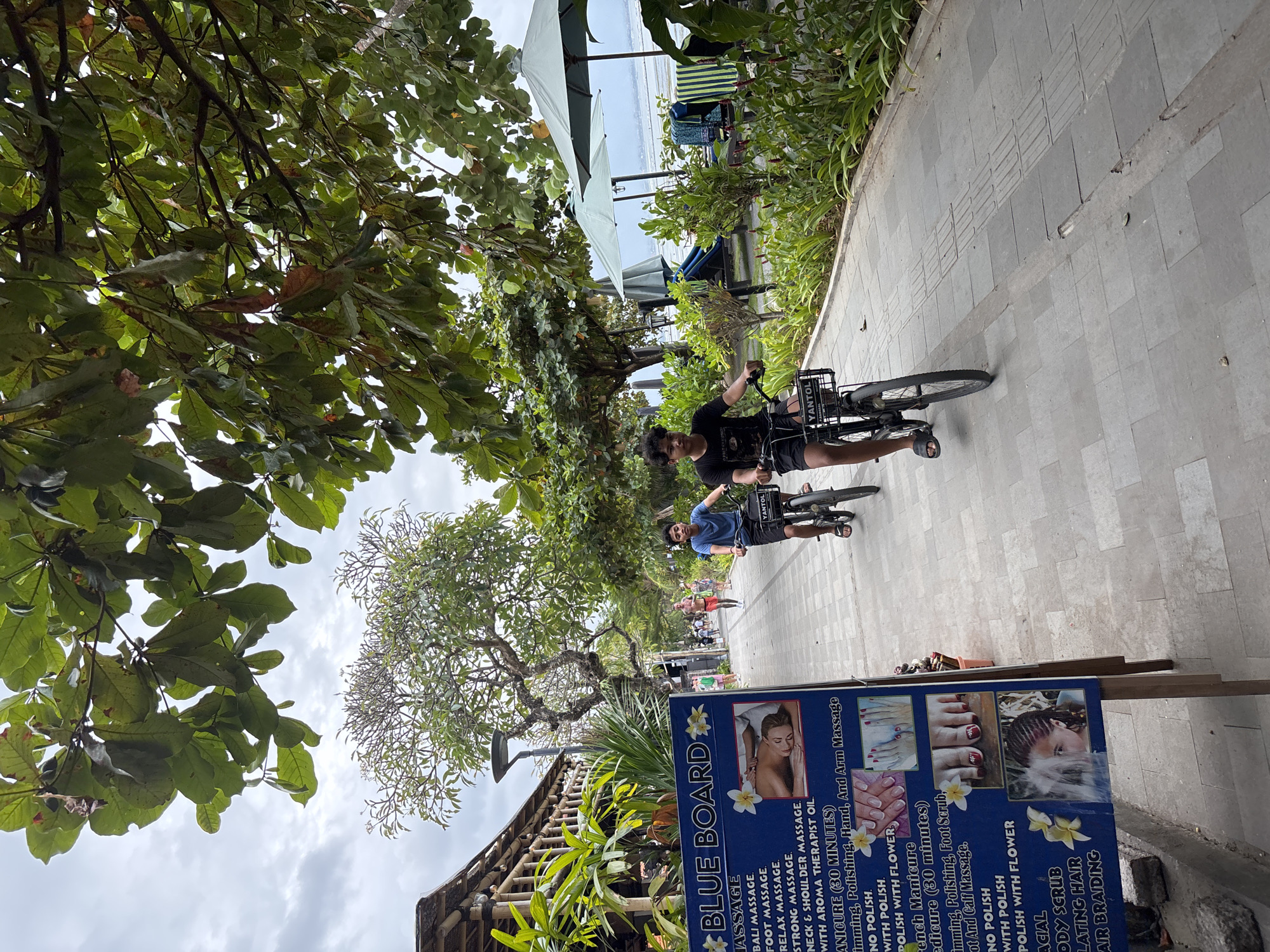 The image depicts a sidewalk scene in Denpasar, Indonesia, likely near a beach area given the presence of palm trees and lounge chairs. In the foreground, a blue sign for "BLUE BOARD" lists various massage and beauty services, including "BALI MASSAGE," "FOOT MASSAGE," "MANICURE (30 MINUTES)," and "HAIR BRAIDING." Two young boys are riding bicycles on the paved sidewalk, which features tactile paving for the visually impaired. The boy on the left is wearing a blue t-shirt, and the boy on the right is wearing a black t-shirt. They are moving away from the camera, towards the background where other pedestrians are visible. The environment includes lush green foliage, trees, and glimpses of the ocean and umbrellas in the distance. The architecture visible on the left suggests a tropical resort or restaurant setting. The sky is overcast.
