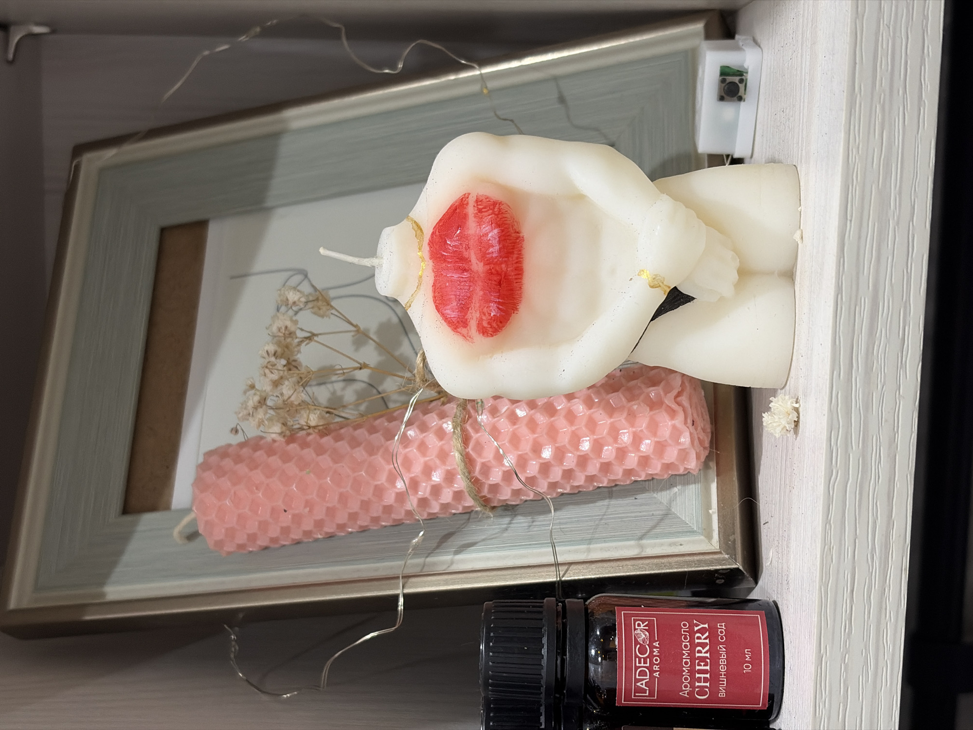A close-up shot depicts various objects on a shelf or in a display setting. Prominently featured is a white, sculpted candle resembling a pair of clasped hands with a red lipstick mark on one of the hands. Adjacent to it is a pink, honeycomb-patterned candle. These are set within a rectangular picture frame that contains dried flowers. In the foreground is a small, dark-colored bottle labeled "Ladecor Aroma Cherry," indicating it is an aroma oil with a cherry scent. The items are positioned close together. A string of small lights is placed near the picture frame. The shot is taken indoors, with a focus on the objects and their arrangement. The location is not discernible from the image, and there is no way to ascertain that this image was taken in Izhevsk, Russia.