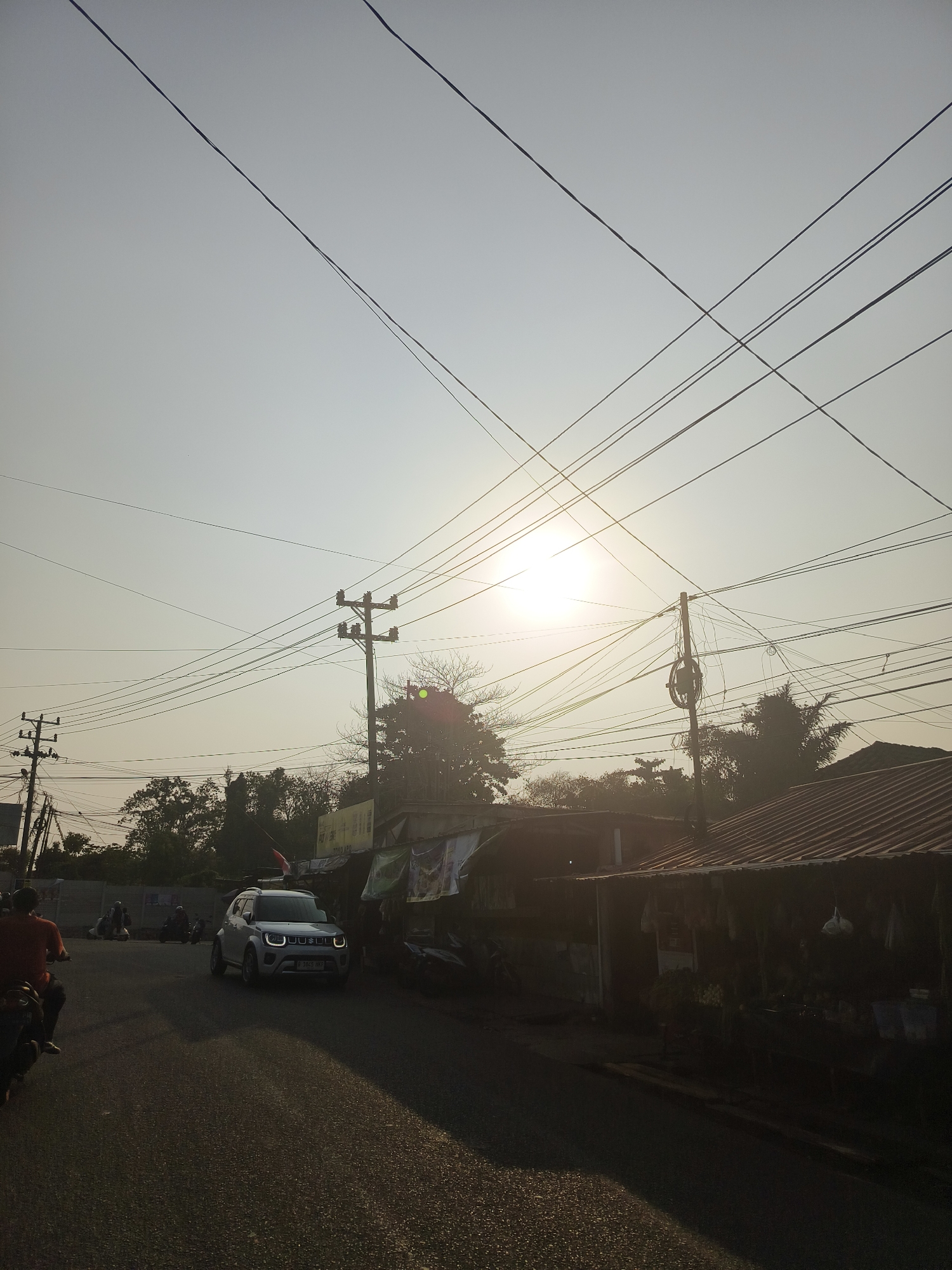 The image depicts a street scene in Pangkalpinang, Indonesia, during the late afternoon or early morning, as indicated by the low angle of the sun casting long shadows. A white Suzuki Ignis car is driving on a paved road, illuminated by the sun. To the right of the road, there are several buildings with corrugated metal roofs and some merchandise displayed outside. Overhead, a complex network of electrical wires crisscrosses the sky, supported by utility poles. In the background, trees and foliage are visible. The sky is bright and hazy, with the sun positioned in the upper center of the frame, creating a lens flare effect. On the left side of the road, a person on a motorcycle is partially visible, with another motorcycle and its rider further down the road. The overall impression is of a typical urban street in a tropical climate with active infrastructure and daily life.