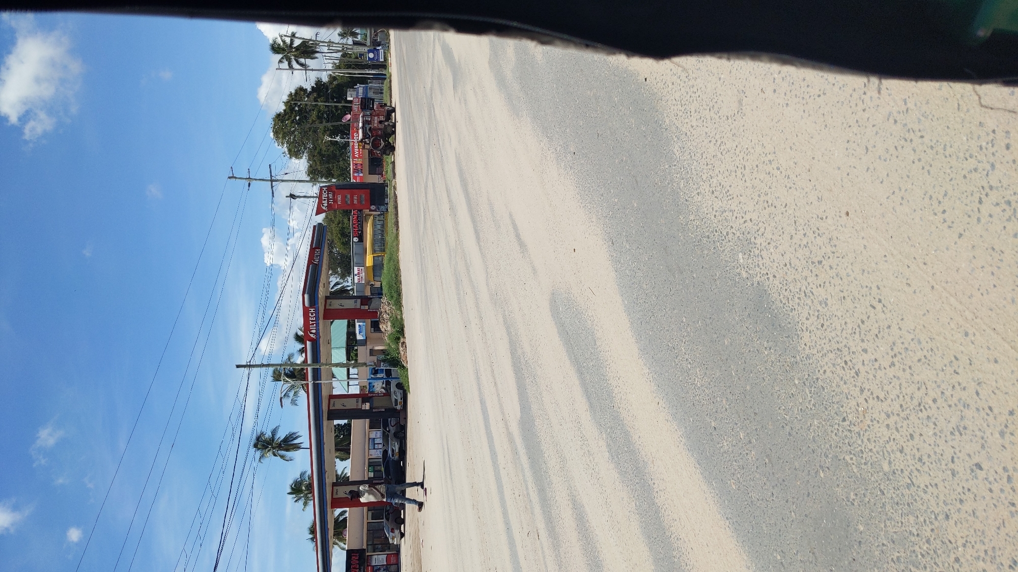 The image shows a view from inside a vehicle, possibly a car, looking out towards a street scene in Dar es-Salaam, Tanzania. On the left side of the street is an OilTech gas station with petrol and diesel fuel pumps. The gas station signage is visible. Palm trees are scattered along the roadside. There are overhead electrical wires and poles. Further down the street, there are various commercial establishments, including what appears to be a tire store. A red tractor and a school bus are also visible. The road itself is paved. A person is standing by the pumps. The sky is blue with some scattered clouds. The asphalt road surface is visible in the foreground, with light and shadows creating texture.