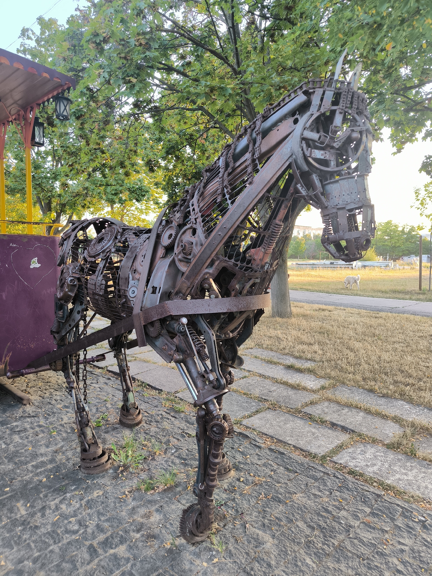 The image displays a metal sculpture of a horse located outdoors. The sculpture is constructed from various metal parts, including gears, chains, and springs, giving it a steampunk aesthetic. The horse sculpture is positioned on a pathway made of stone slabs, with dry grass and trees visible in the background. A light-colored dog is seen walking on a leash in the distance. The scene suggests a public park or outdoor art installation. The location is identified as the Alley of Classics in Chișinău, Moldova. The time of day appears to be late afternoon or early evening, given the warm lighting.