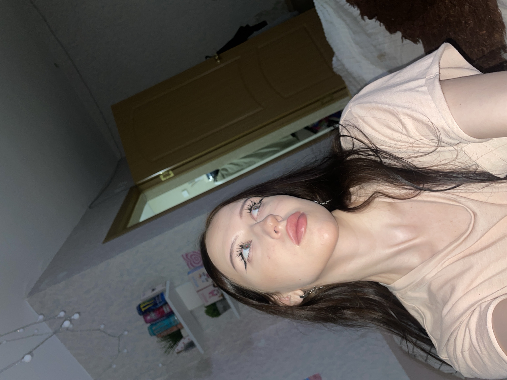 The image shows a young woman with long, dark hair and light skin, wearing a light peach-colored t-shirt. She has dramatic black eyeliner and light pink lipstick. She is looking upwards and to the right. In the background, there is a white shelf with several items, including what appear to be cans of energy drinks and small potted plants. A string of fairy lights with small white bulbs hangs on the wall above the shelf. Further back, a wooden door is visible, partially ajar, revealing a glimpse of what might be a closet or another room. The setting appears to be a bedroom with wallpaper and a textured wall. The lighting is dim, suggesting it is nighttime or the room is not well-lit. The image quality is typical of a smartphone camera. There is no specific information in the image that would allow for identification of the location within Votkinsk, Russia.