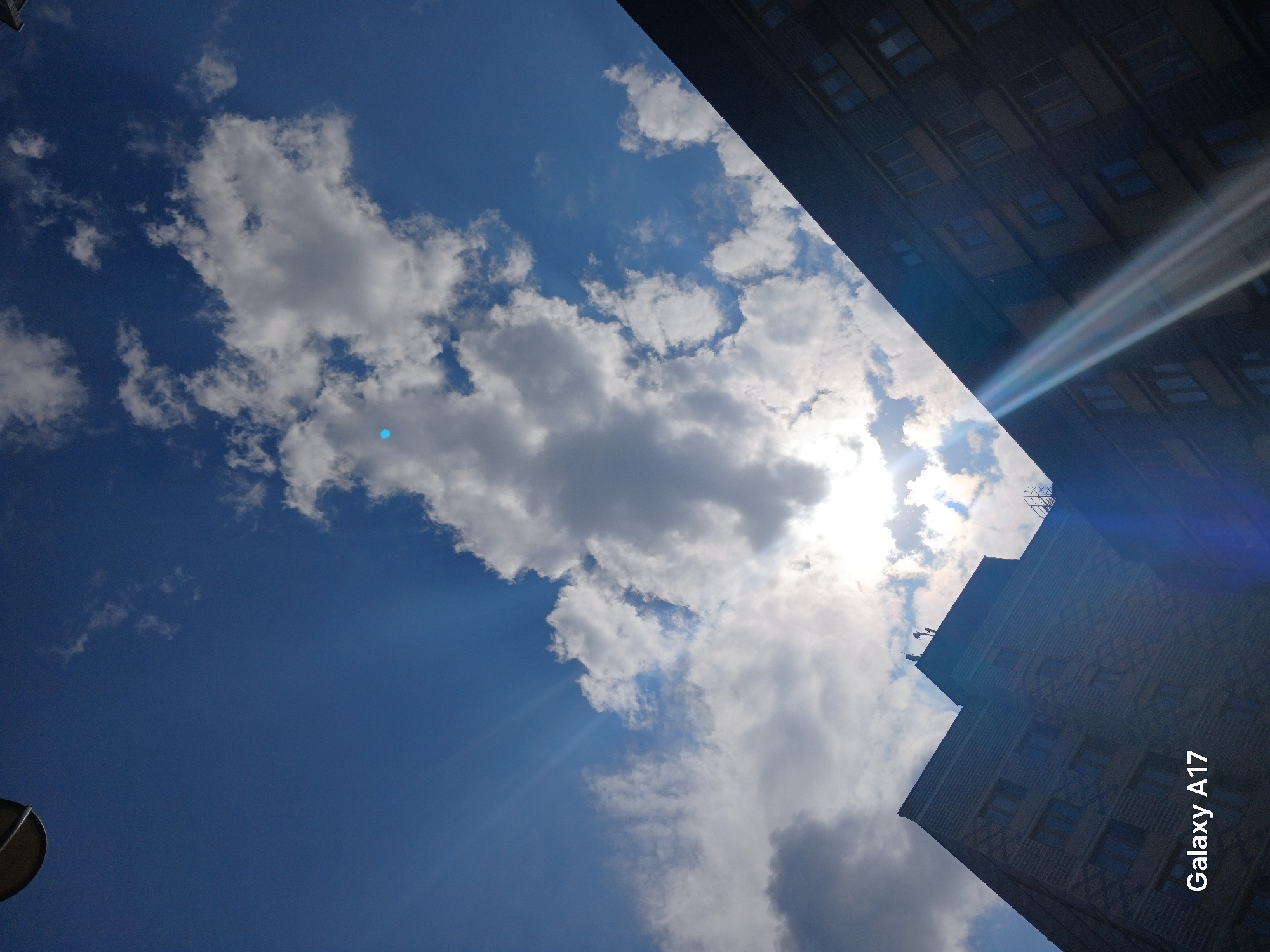 The image, taken with a Galaxy A17, shows a view upward into a bright, partially cloudy sky next to a building. The lower-left corner includes a gray circular object, likely part of a traffic or street light. The majority of the image captures the blue sky with varying cloud formations from small, scattered white clouds to larger, denser gray clouds. The sun is partially obscured by clouds, resulting in light rays extending downward. The building, likely located in an urban area of Yekaterinburg, Russia, takes up the upper-right section of the frame. It has multiple windows and appears to be made of brick. The building's perspective indicates the photo was taken from a low angle.