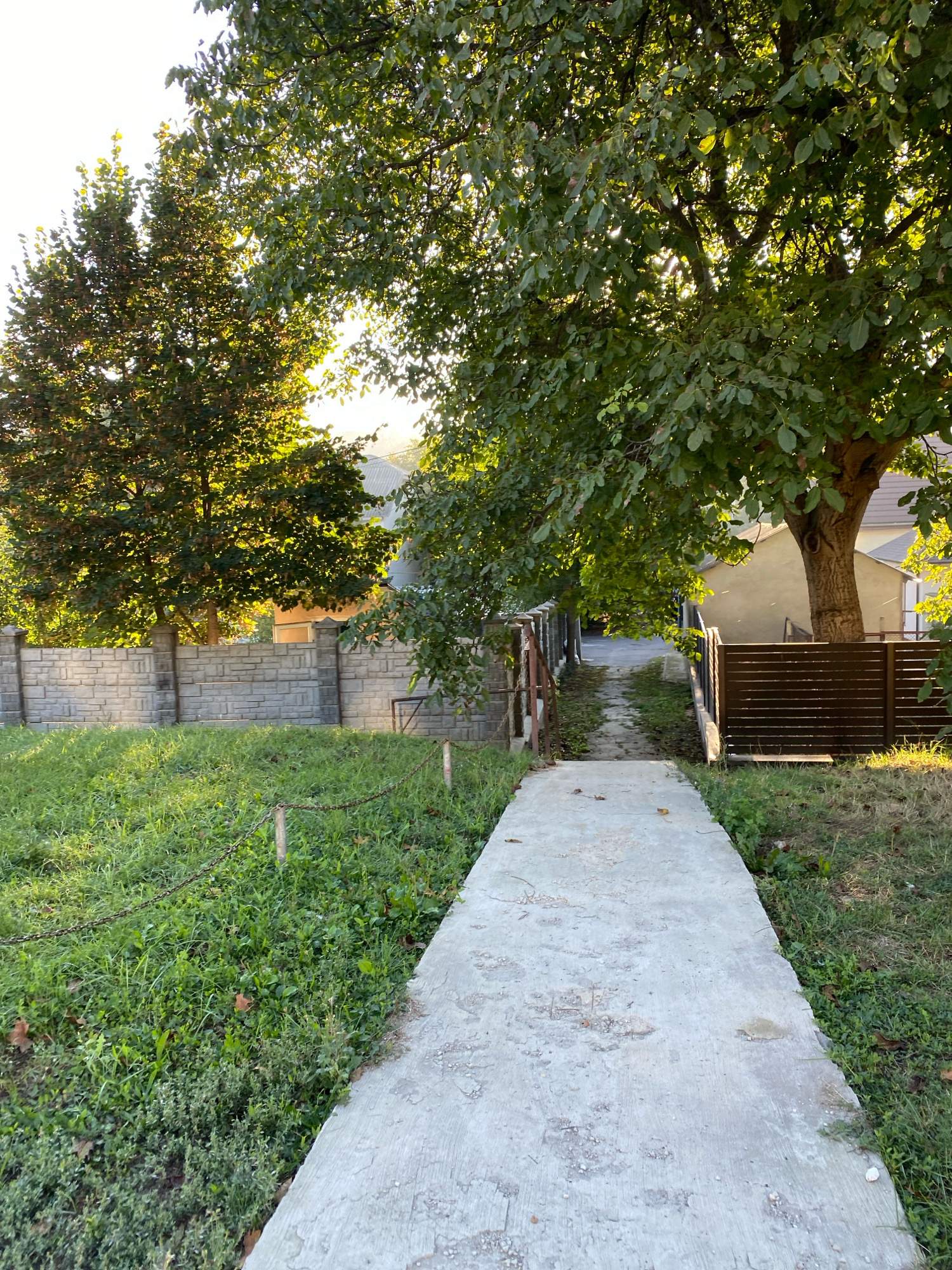 The image depicts a concrete pathway leading through a yard with abundant green grass and trees. A light-colored stone fence lines the left side of the pathway, with a chain fence in the foreground. On the right, a dark horizontal slatted fence runs alongside a portion of the pathway, partially obscured by the leaves of a large tree. The pathway itself appears aged, with some visible wear and tear. In the background, a house with a light-colored facade is visible, peeking through the foliage. The scene is bathed in the warm glow of sunlight, suggesting it is either early morning or late afternoon. The overall impression is of a tranquil, residential outdoor space. The image does not contain any people or identifiable landmarks specific to Chișinău, Moldova.