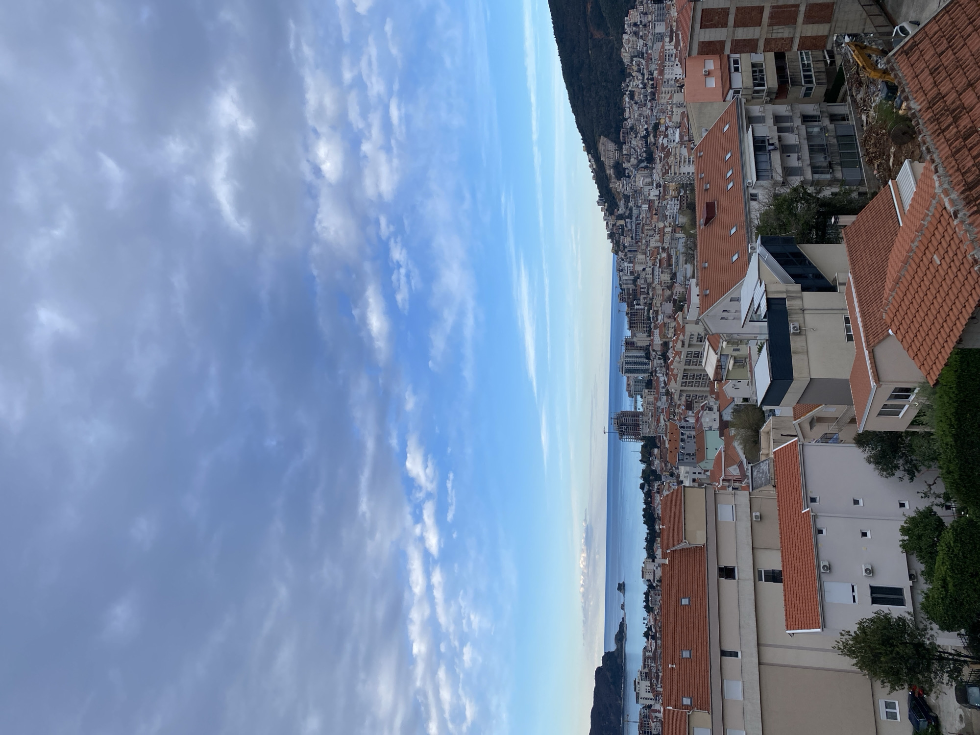 The image presents a cityscape with a body of water (likely the Adriatic Sea) bisecting the view horizontally. The sky is a mix of light blue with scattered clouds on the left, transitioning to a brighter blue towards the sea.
On the right, a densely packed urban area with buildings of varying heights and architectural styles is visible. Predominantly, the buildings feature red-tiled roofs. Modern apartment blocks mix with older structures. A portion of a green, wooded hillside rises in the background above the buildings. The location is Budva, Montenegro, as indicated by the recognizable characteristics of its architecture and coastline. There are no people visible in the image.