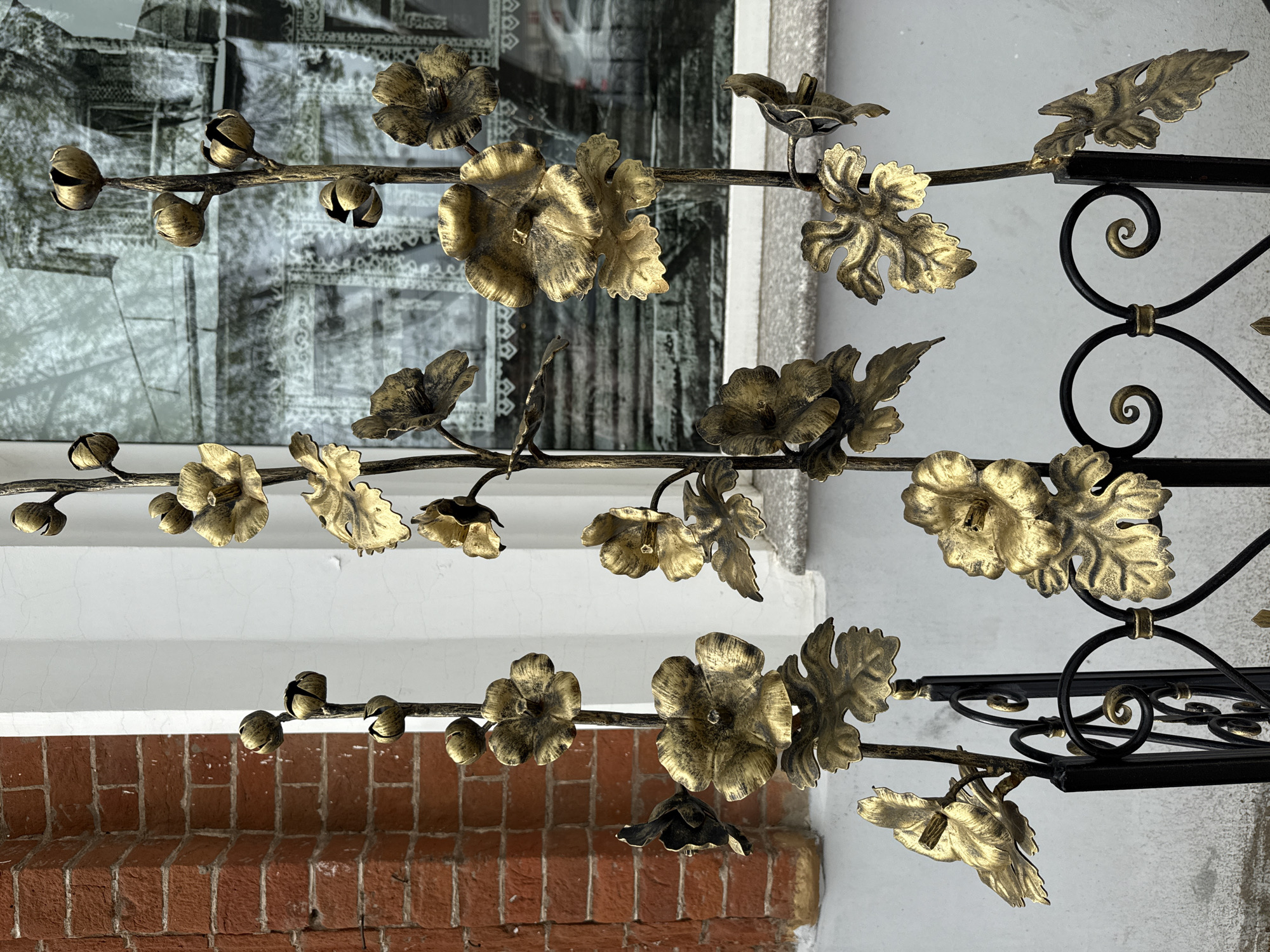 The image shows a decorative iron fence featuring golden metal flowers and leaves. The fence is positioned in front of a building, evident by the brick wall at the bottom, the white wall above, and the window reflecting an elaborate interior structure in the background. The wrought-iron elements of the fence include floral motifs and decorative scrollwork. The location is Votkinsk, Russia, based on visual elements.