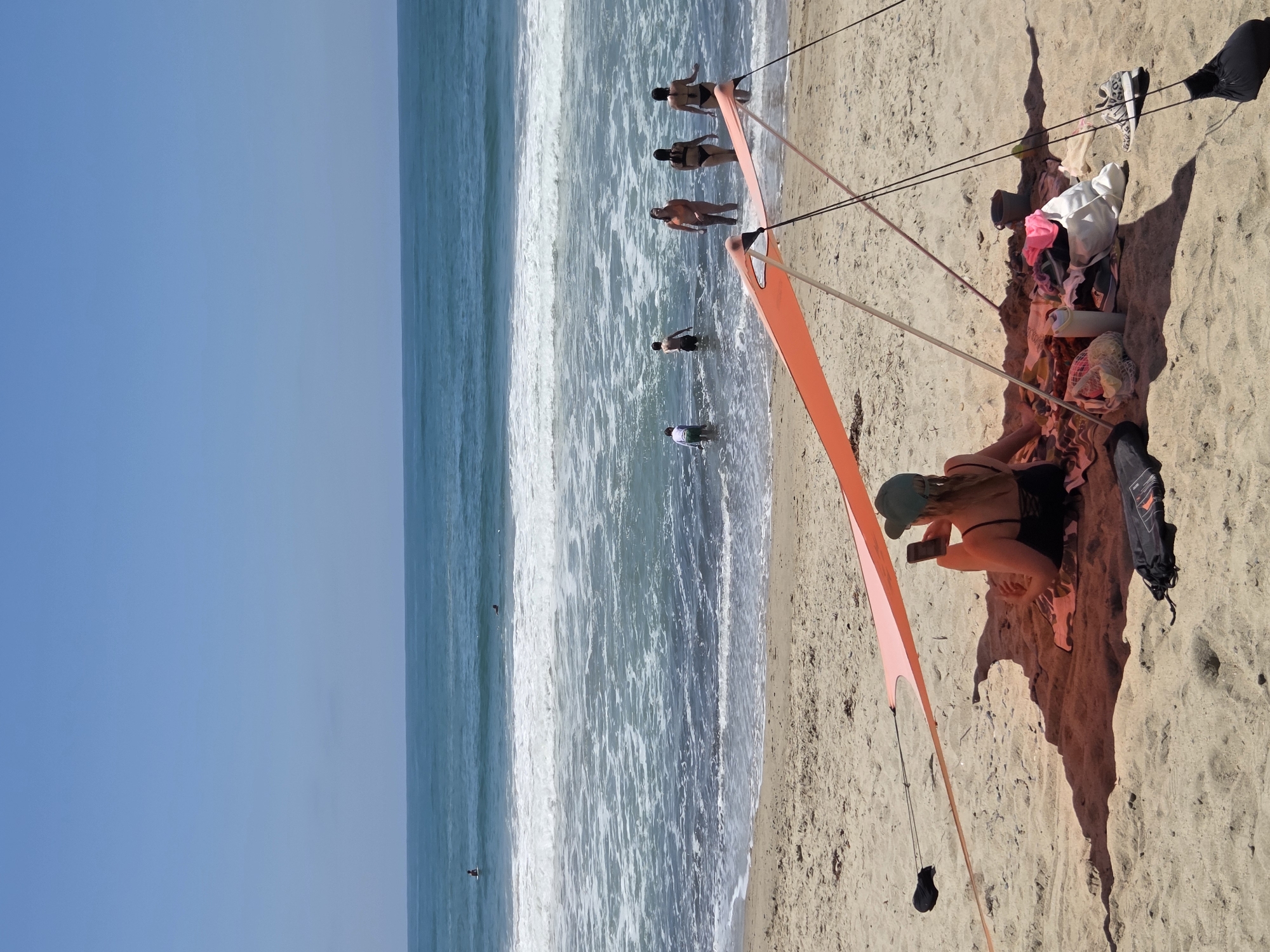 The image depicts a sunny day at a beach, likely in San Clemente, California, with the Pacific Ocean in the background. In the foreground, a person is sitting on a towel under a small orange beach tent, looking at their phone. Scattered around them are beach accessories including a water bottle, a bag of items, and a pair of sneakers. Further out in the ocean, several people are visible wading in the water and some are in the process of surfing. The sand is light brown, and the water is a clear blue with white waves. The sky is a bright, cloudless blue.