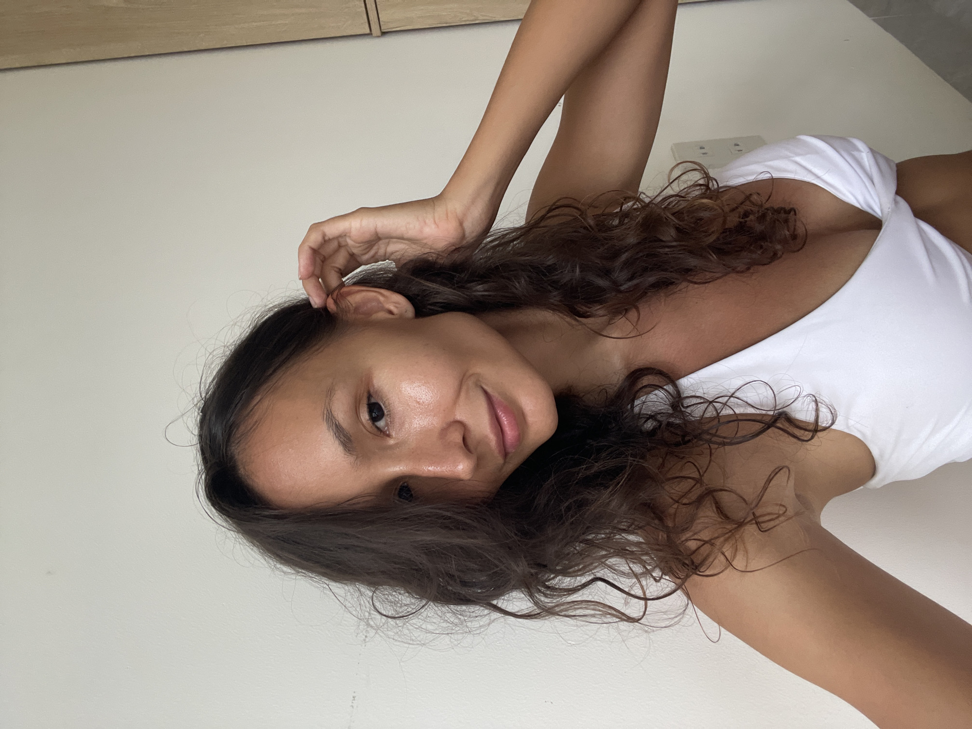 A medium-shot selfie features a young woman with long, dark, wavy hair and light brown skin. She is wearing a white bikini top with a V-neckline. Her left hand is raised to her head, tucking her hair behind her ear. Her right arm is bent and resting against her side. She is looking directly at the camera with a slight smile and her eyes are open. Her skin appears dewy and slightly shiny. The background is a plain white wall with a wooden cabinet on the right. The scene is indoors, likely in a residential setting, with no indication of being in Phu Thuy Ward, Vietnam.