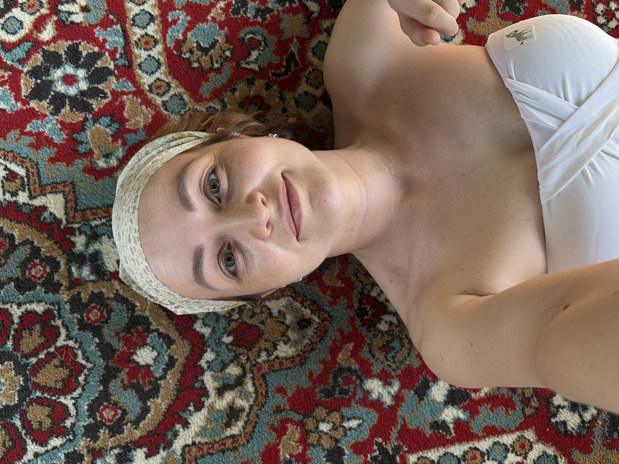 A woman lies on an ornate patterned rug, likely indoors. She wears a white strapless top and a patterned headband. Her expression is neutral with a slight smile. There is no information to indicate this photo was taken in Chaikovsky, Russia.