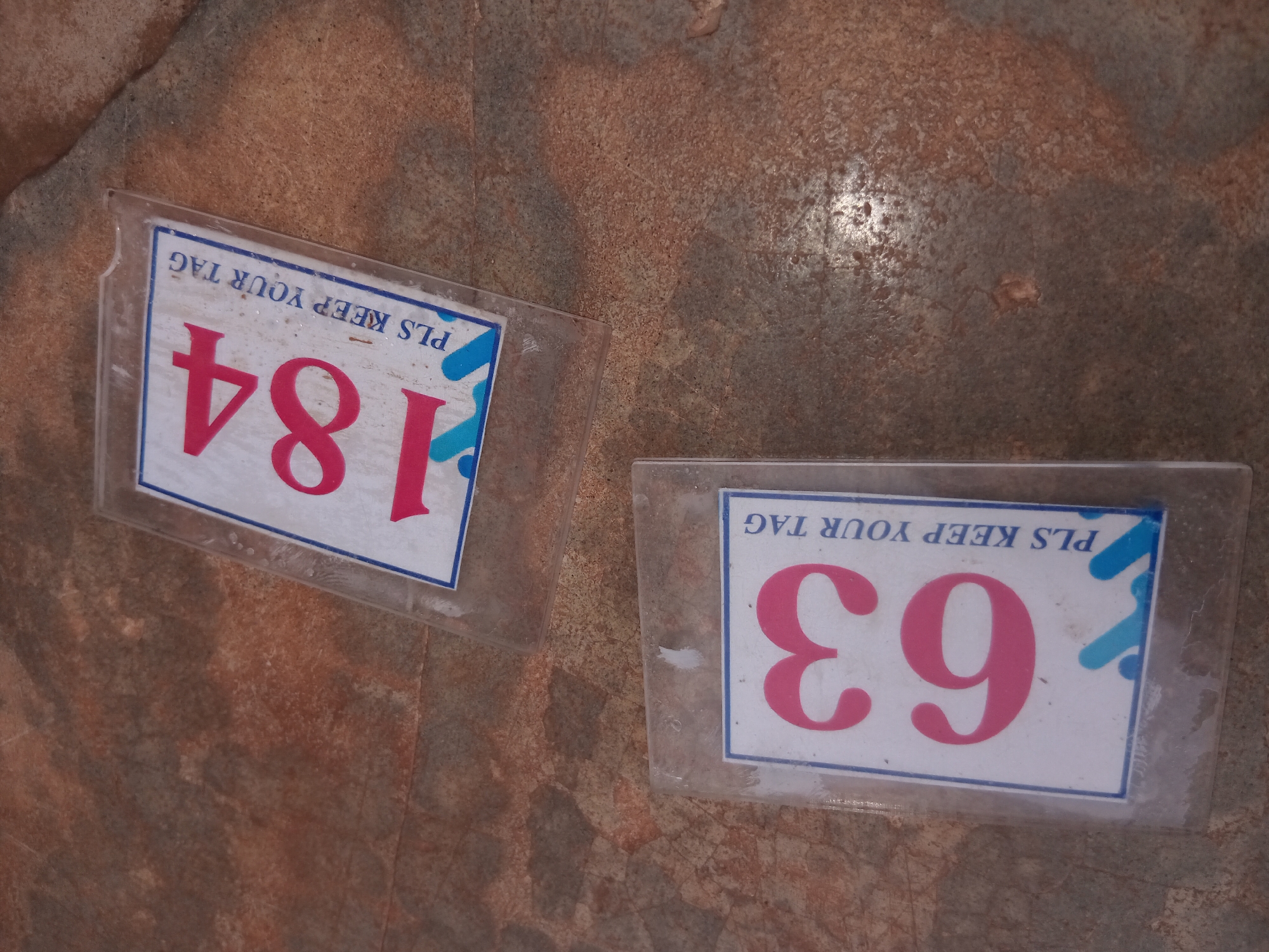 Two plastic tags are on a textured brown surface. The tag on the left displays the number "184" in pink. The tag on the right displays the number "63" in pink. Both tags have "PLS KEEP YOUR TAG" printed above the numbers. The background is a textured surface with variations of brown and grey tones. There is nothing to indicate this photo was taken in Shagamu, Nigeria.