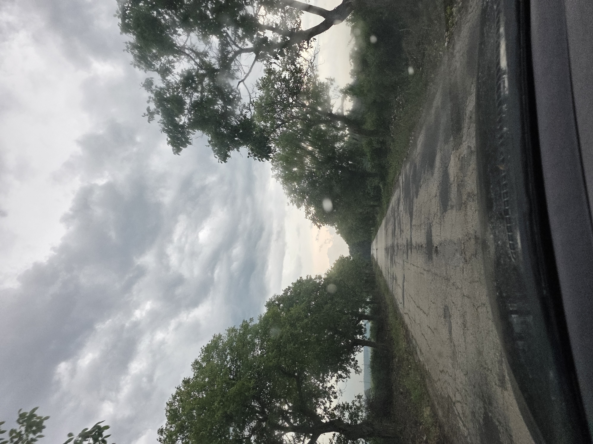 The image captures a rural road in Kamenar, Bulgaria, viewed from inside a vehicle. The scene is dominated by a long, wet, and worn asphalt road that recedes into the distance, flanked on both sides by dense, leafy green trees. These trees form a natural avenue, with their canopies partially obscuring the sky overhead.
The sky is heavily overcast with thick, grey clouds, suggesting recent or ongoing rain. This is further supported by the visibly wet surface of the road and numerous water droplets clinging to what appears to be the car's side window or lens, seen especially along the right edge of the frame. Despite the widespread cloud cover, a brighter, lighter area is discernible on the horizon, hinting at the sun trying to break through the clouds or reflecting off a distant body of water. Sunlight also filters through the tree leaves in some areas, creating subtle highlights.
No people are visible, and the implied activity is travel along this road. The dark frame of a car door or window is prominent on the right side of the image, defining the viewer's perspective. The overall impression is one of a serene, possibly damp, journey through a tree-lined landscape under a dramatic cloudy sky. There is no visible text in the image.