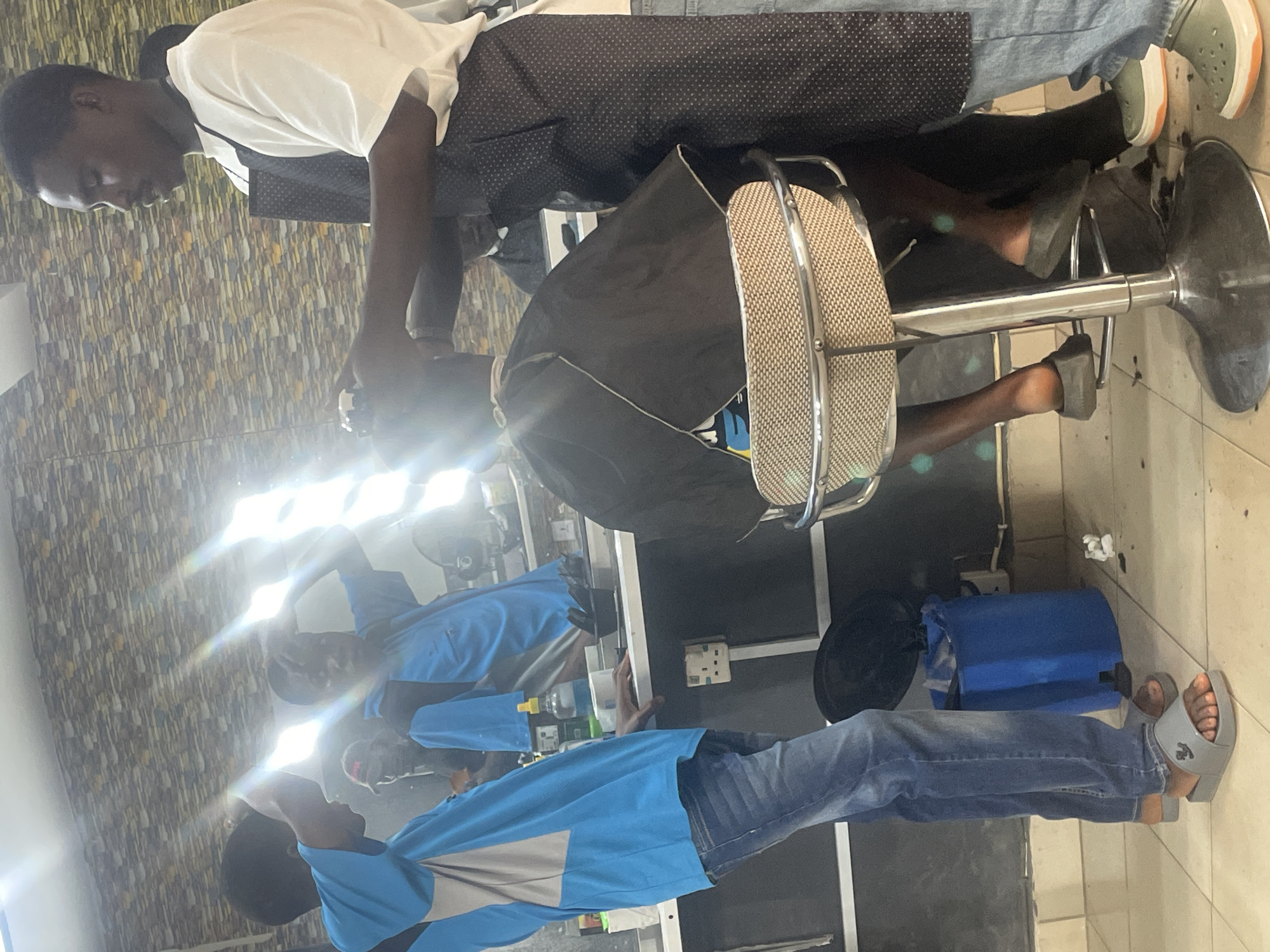 A barber is giving a haircut to a client in a shop. The barber is wearing a white shirt and black apron. The client is seated in a barber's chair, draped with a black cape. Other individuals are present in the background. The location is in Jos, Nigeria.
